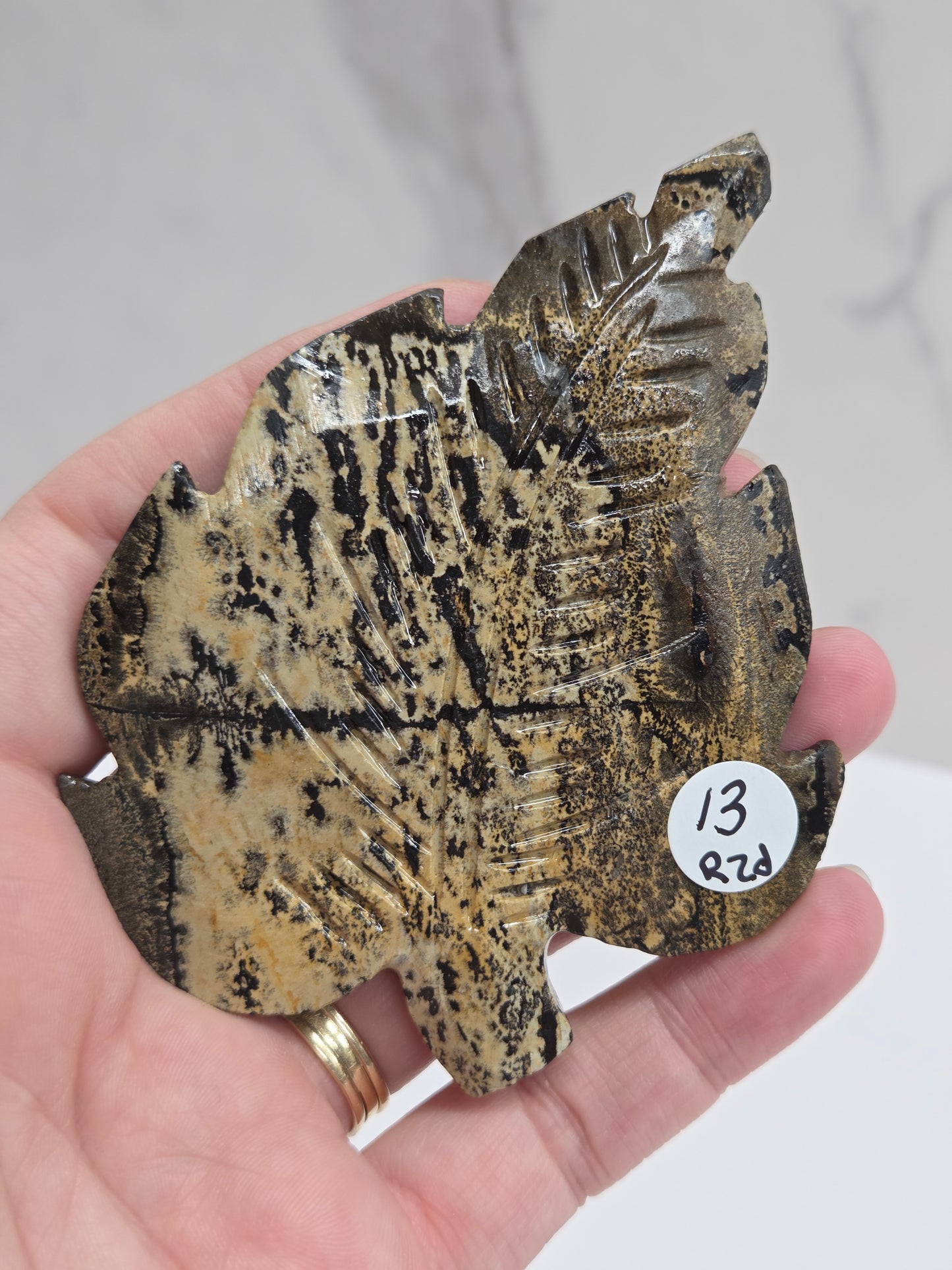 Hand holding a Picture Jasper leaf carving with black, gold, and white patterns against a neutral background