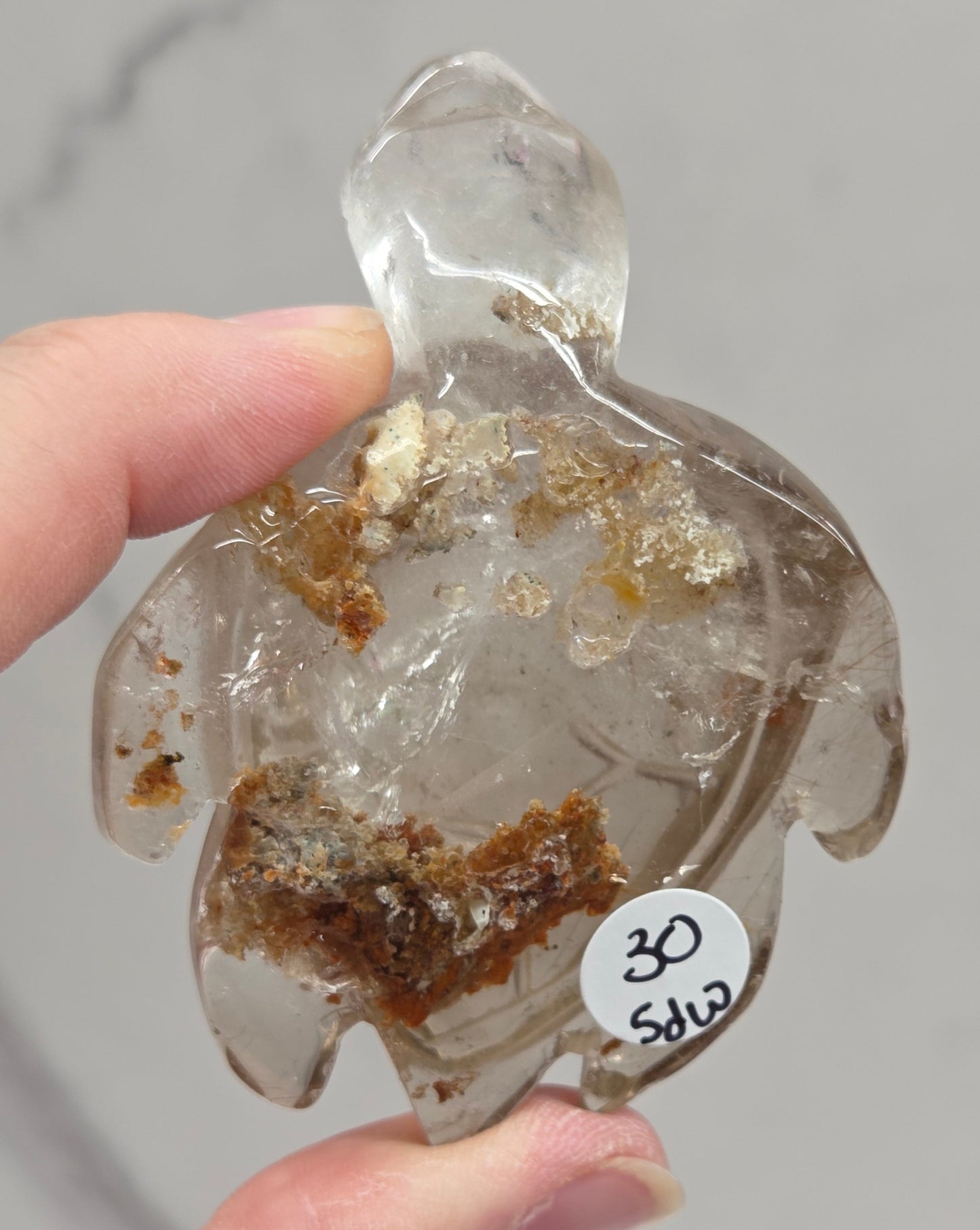 Clear crystal with embedded brown material held by a hand against a neutral background