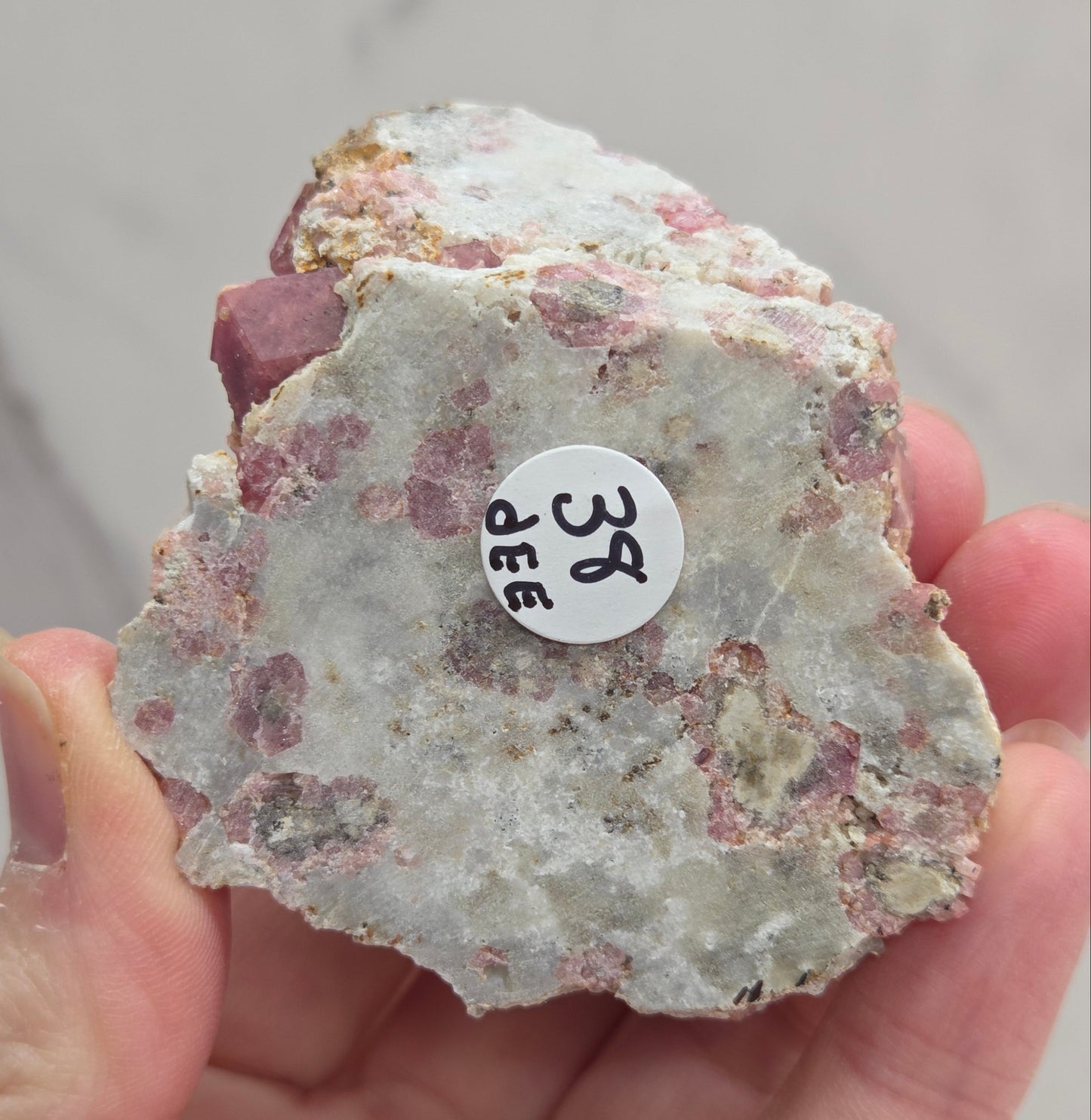 Hand holding a rough mineral specimen with a label on a neutral background