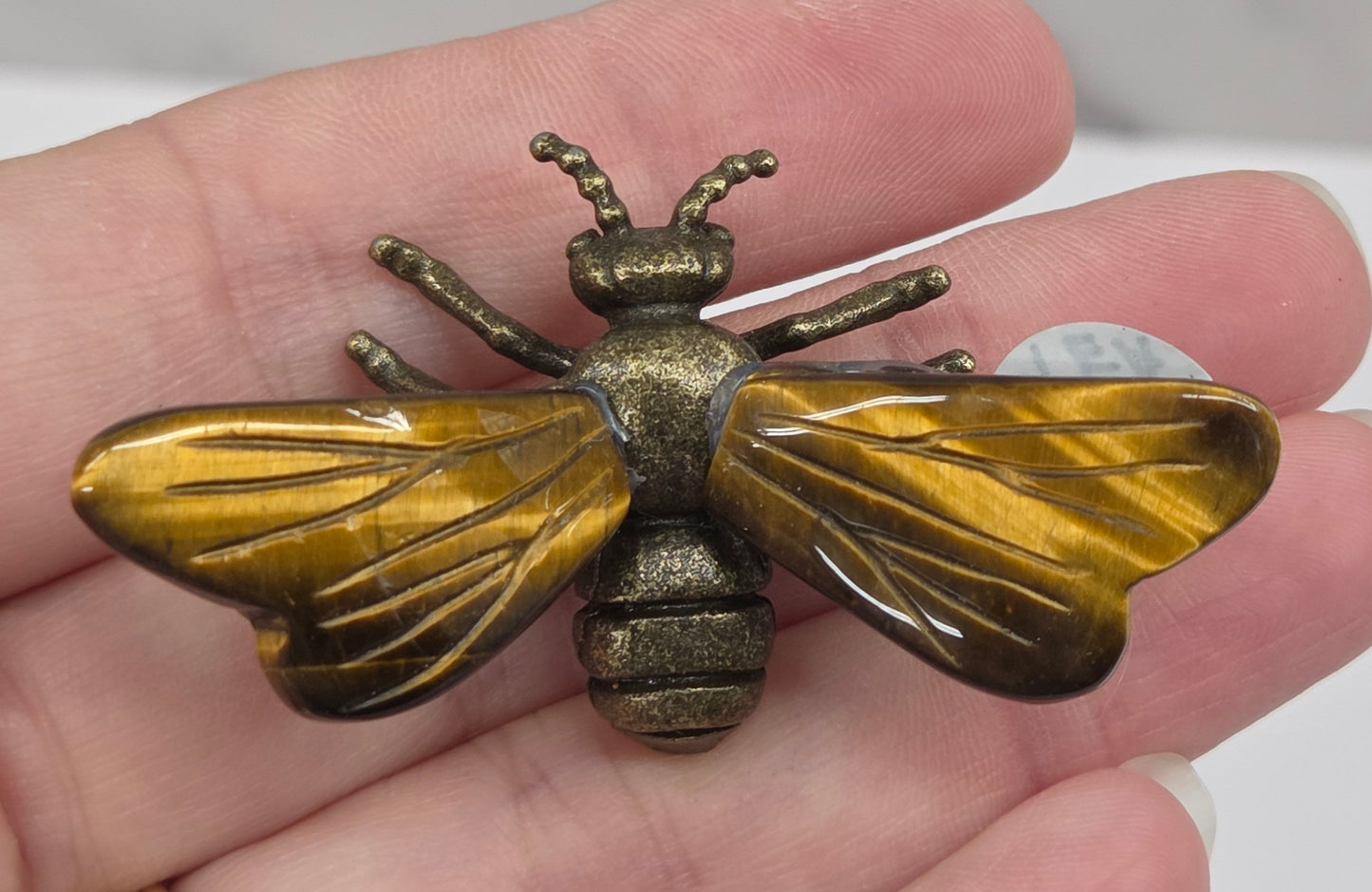 A close-up of a hand holding a vintage-style brass bumblebee brooch. The brooch features detailed wing and body textures with a warm gold and bronze metallic finish.