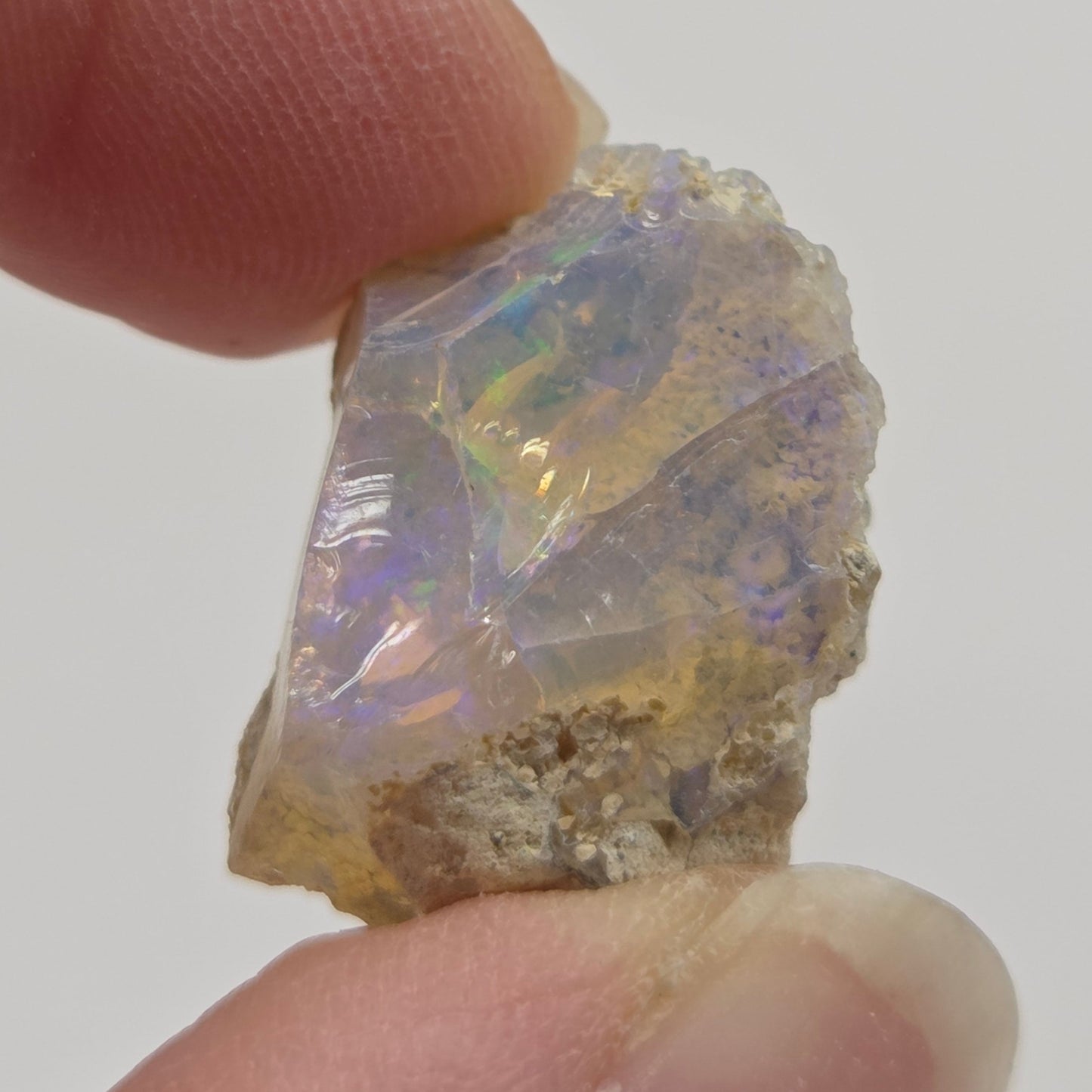 A raw, rough opalite crystal held between two human fingers to show its size and texture. The opalite displays a milky white base with vibrant, shifting iridescent flashes of blue, green, and purple.