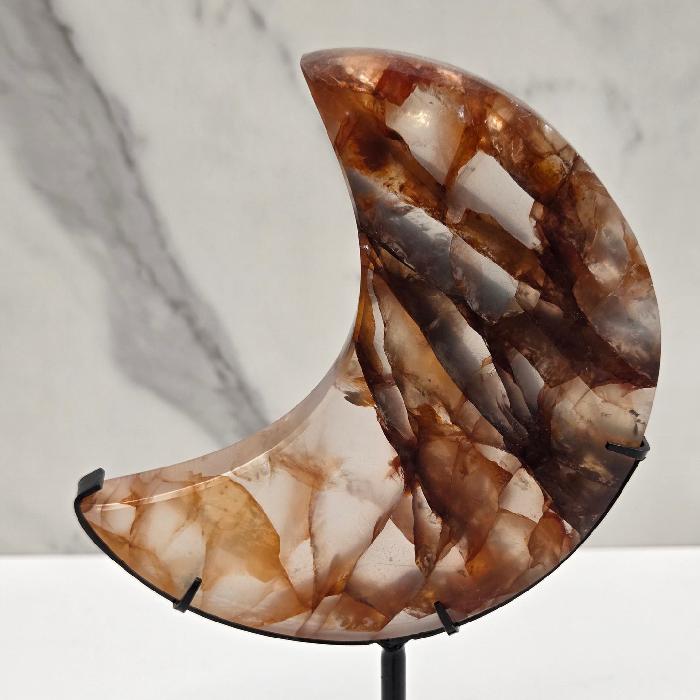 A close-up of a crescent-shaped wall sconce with a polished brown and cream agate stone shade, mounted on a black metal rod against a white marble and white wall background.