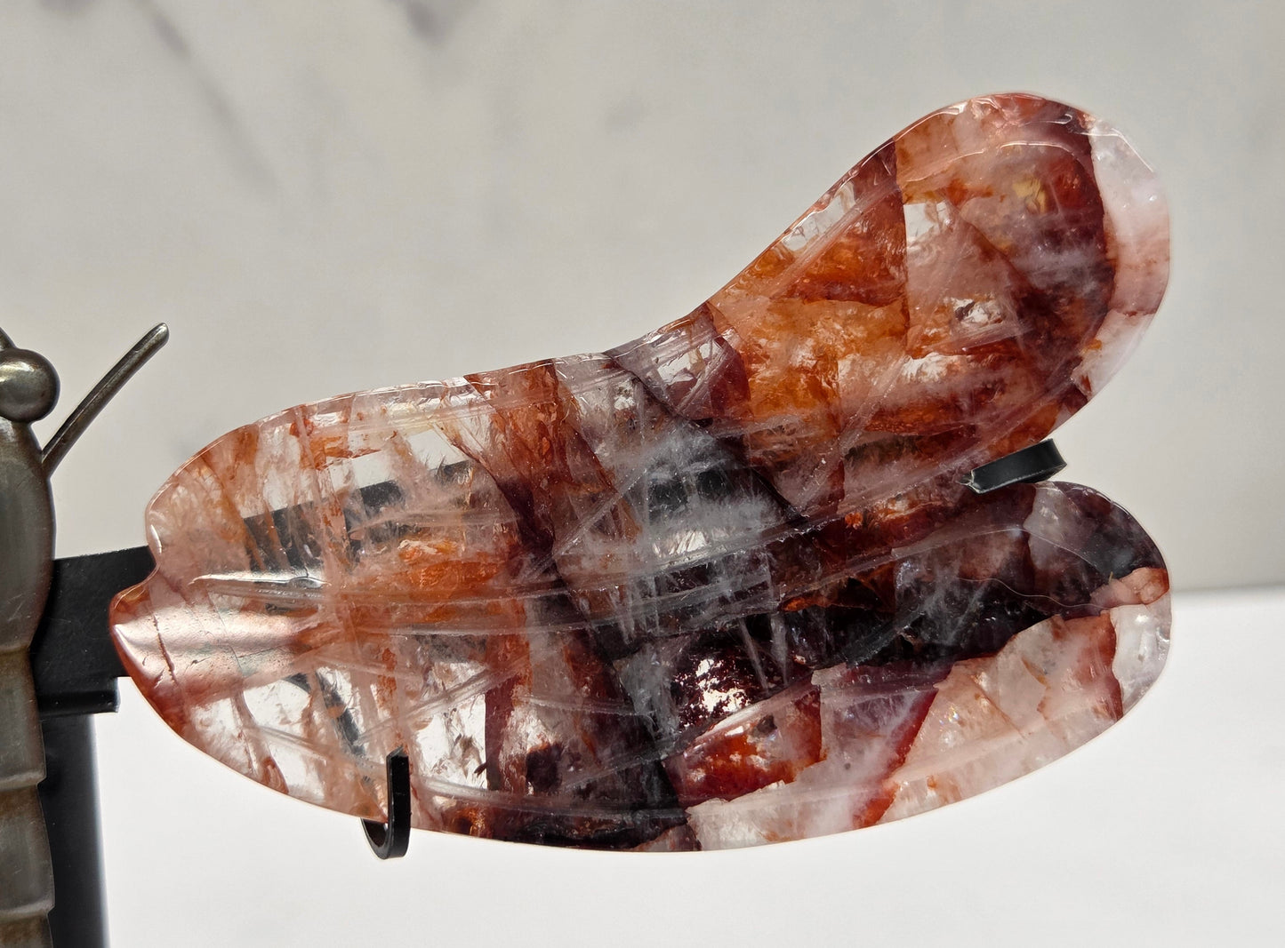 A close-up of a dragonfly sculpture made from polished translucent brown and smoky amber crystals mounted on a dark metal stand, set against a light gray marble-patterned background.