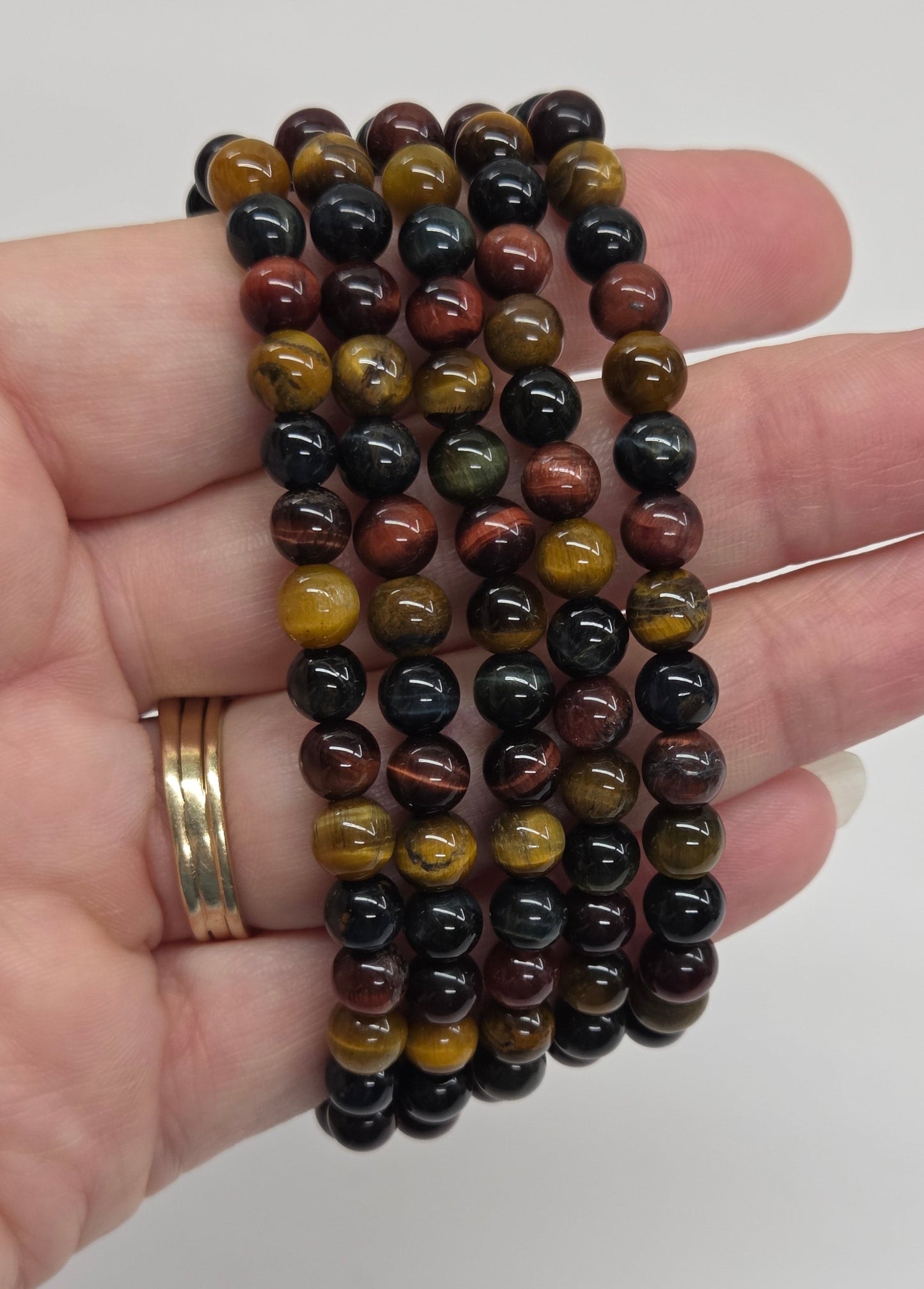 A hand holding three beaded bracelets made of polished round stones in black, brown, and golden-yellow tiger-eye patterns, with a gold ring on the finger, against a plain light gray background.