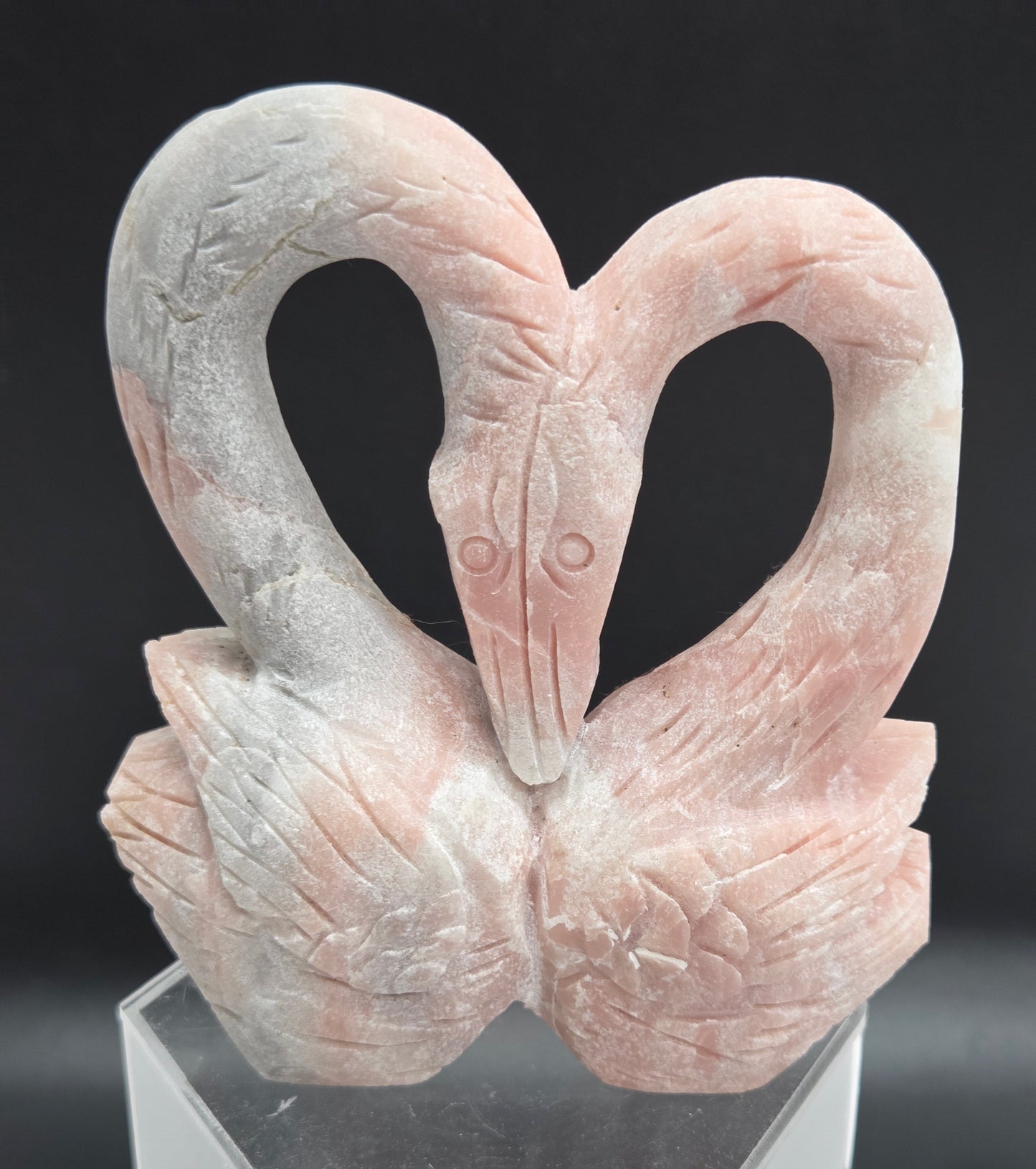 A close-up of a sculpted decorative object featuring two intertwined looped forms resembling a stylized bird or abstract sculpture, mounted on a clear acrylic stand against a dark background.