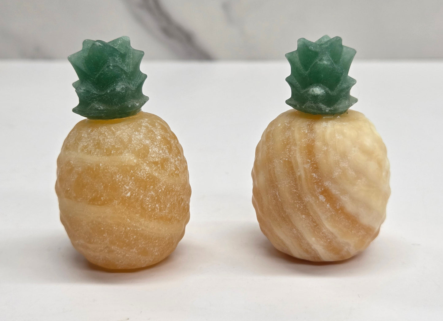 Two small pineapple-shaped decorative objects with textured pale yellow bodies and green tops, standing on a white surface against a light gray marble-patterned background.
