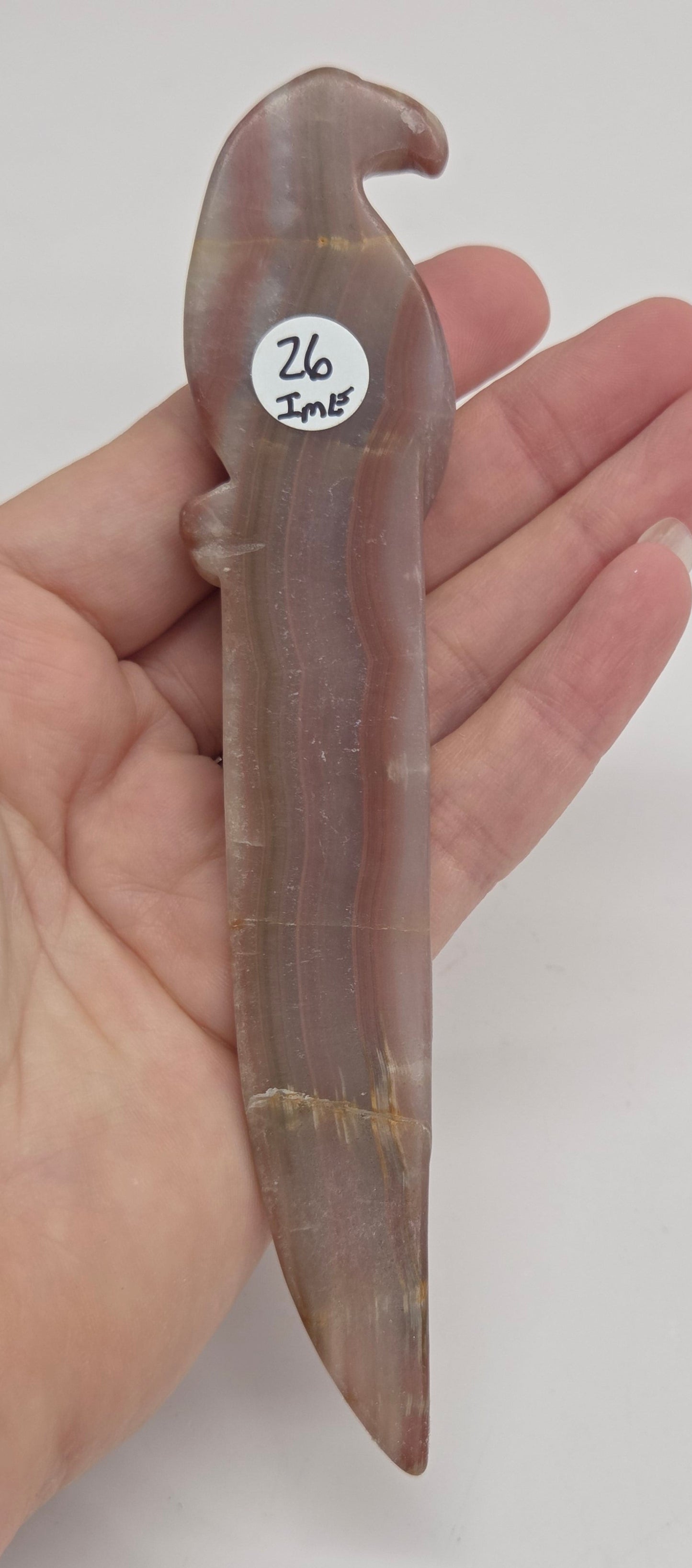 A hand holding a small carved stone object, likely a miniature tool or charm, made of a translucent brownish-gray stone with a small white sticker labeled '26 Jazzy' on the side.