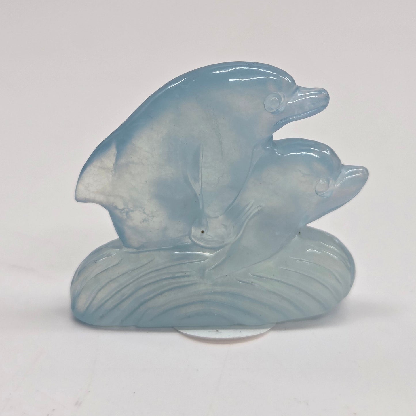 A small carved light blue stone figurine depicting two dolphins on a wave, displayed on a clear stand against a plain white background.