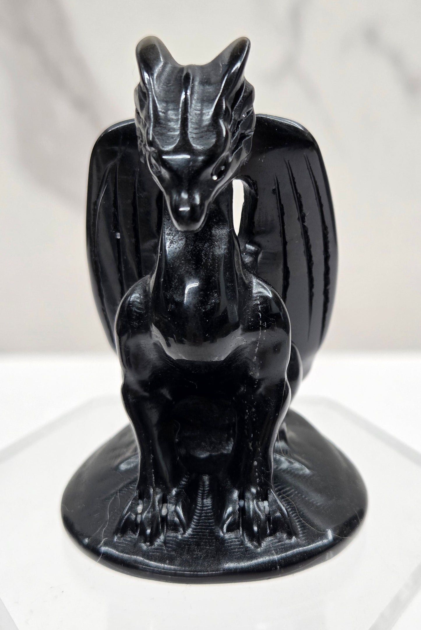 A glossy black ceramic dragon figurine mounted on a clear acrylic hexagonal display stand against a white marble-patterned background.