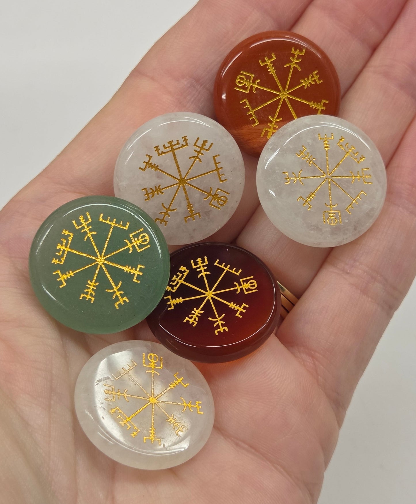 Six round cabochon gemstone pendants with gold Norse runes and compass motifs, held in a person's hand to show scale.
