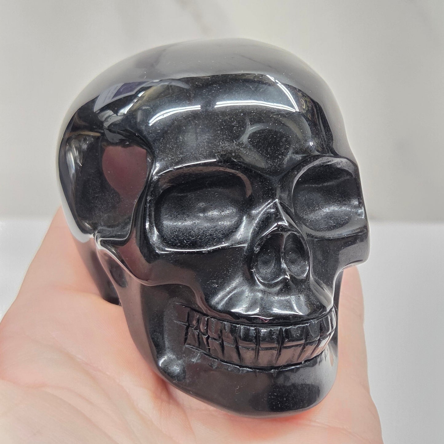 A glossy black skull-shaped object being held against a light gray marble-like background, with a human hand visible on the right side.