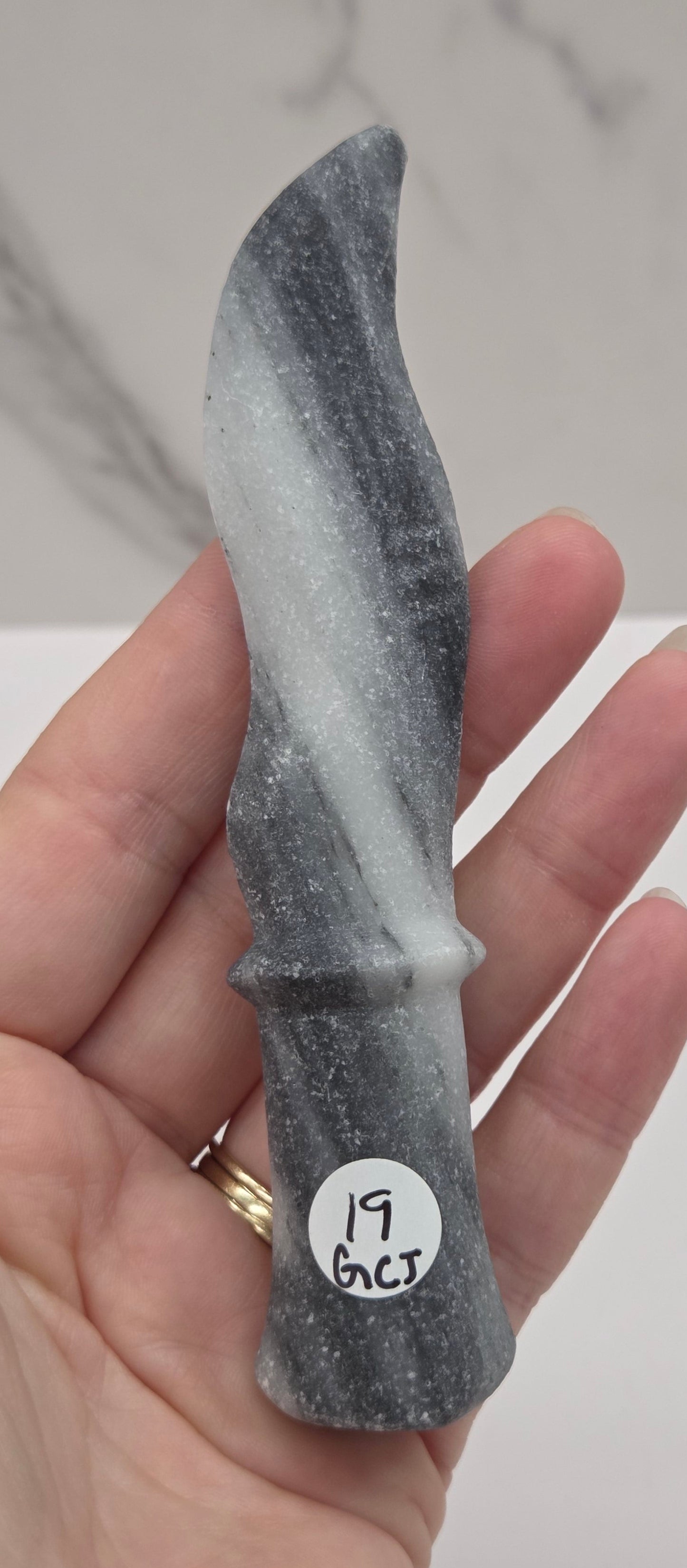 A hand holding a small gray and white marbled stone knife blade with a circular white sticker that reads 'JL 6.5' on a light background.