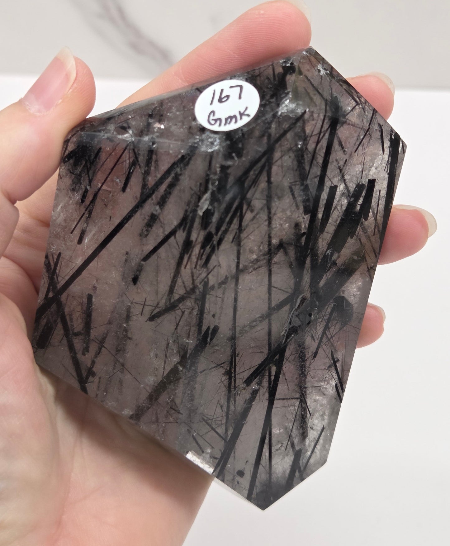 A hand holding a faceted clear crystal with dense black needle-like inclusions, likely a rutilated gemstone. A small white sticker on the crystal reads '167 6mk'.