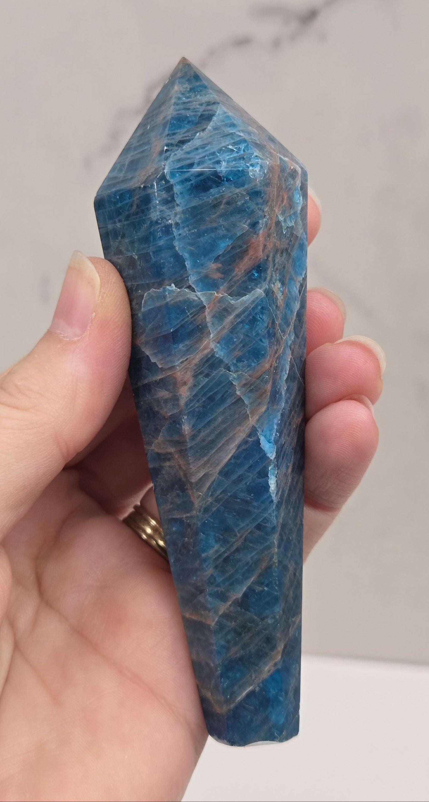 A hand holding a polished blue apatite crystal point against a light marble background.
