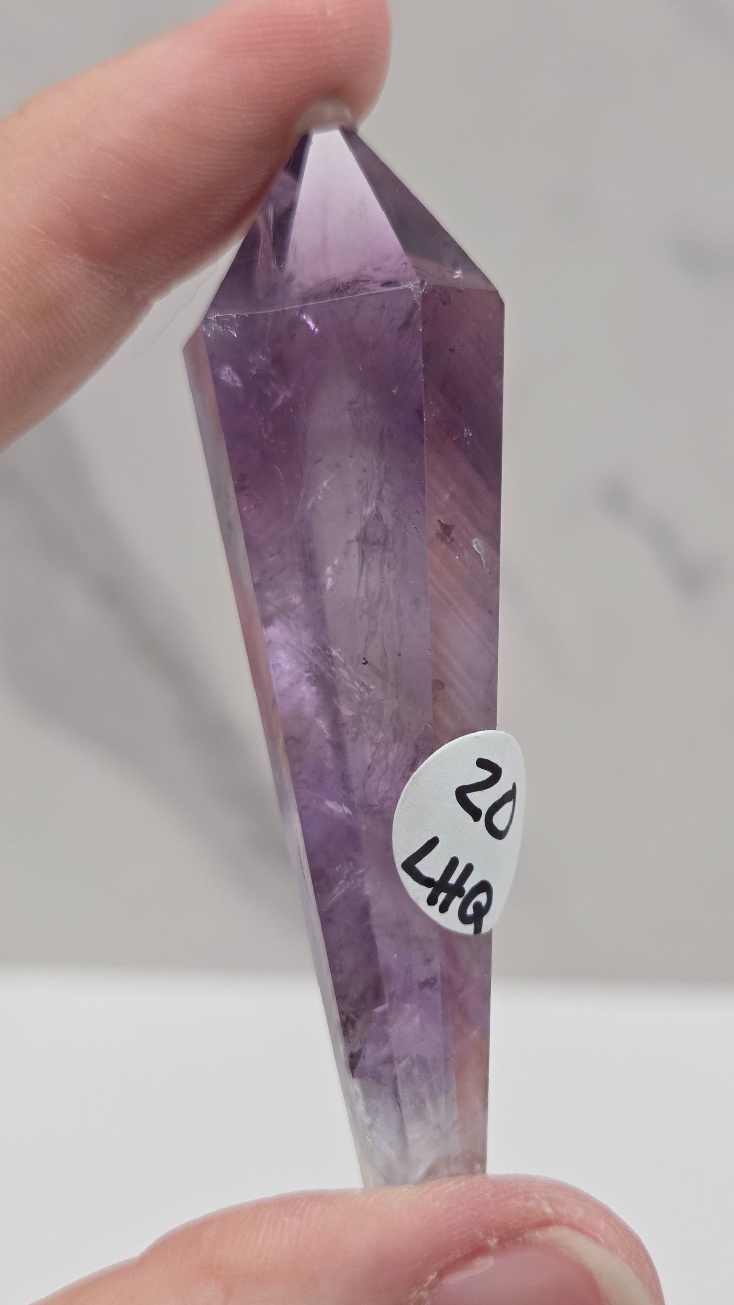 A hand holding a faceted purple crystal point, likely amethyst, with a small white sticker labeled '22 847' attached near the center. The background is a soft-focus light gray marble-like surface.