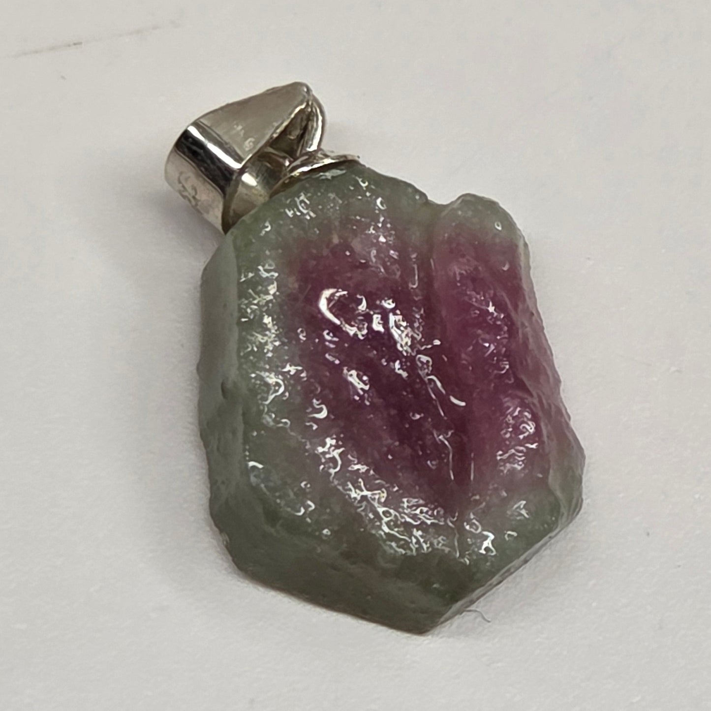 A raw, rough-cut gemstone pendant with a silver bail, showing a distinct color transition from pinkish-purple on one side to green-gray on the other, photographed on a plain light background.