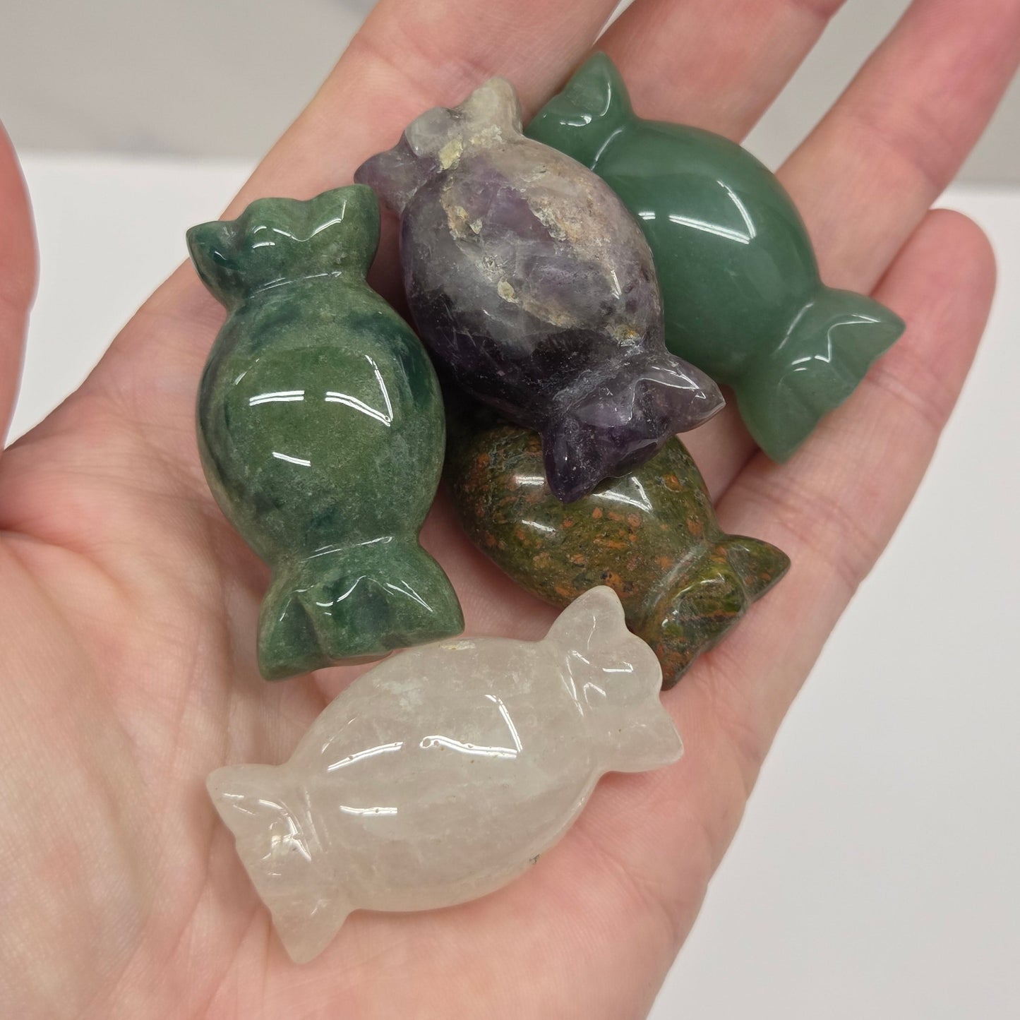 Five small polished stone figurines shaped like wrapped candies are displayed on an open hand. The set includes green, purple, olive green, and milky white stones with a glossy finish.