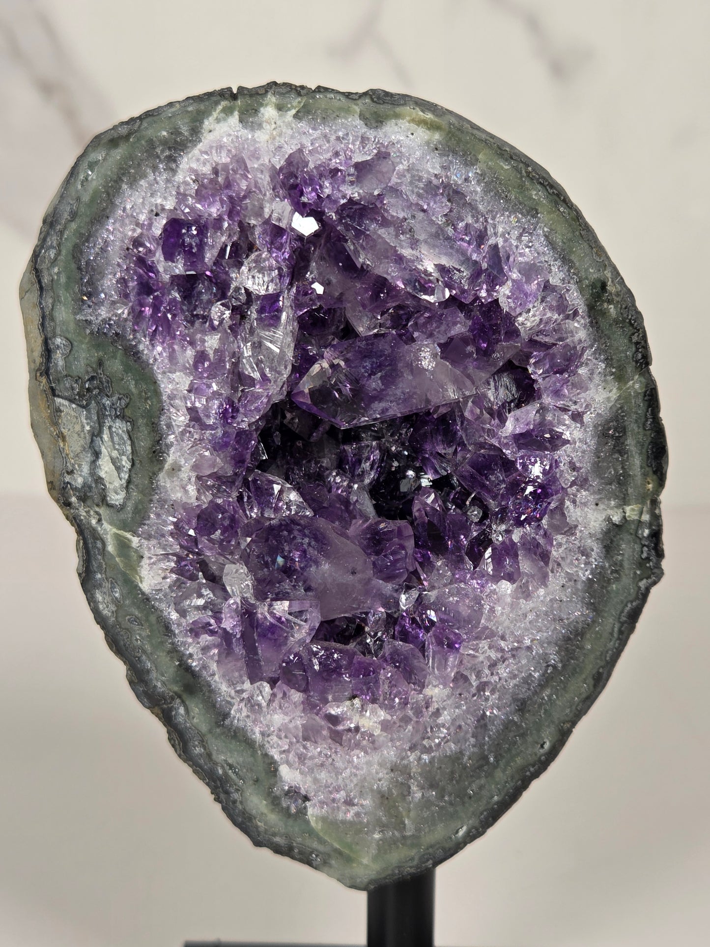 A raw amethyst geode specimen with a dark gray outer matrix and a dense cluster of purple crystals inside, photographed close-up on a light marble-like surface.