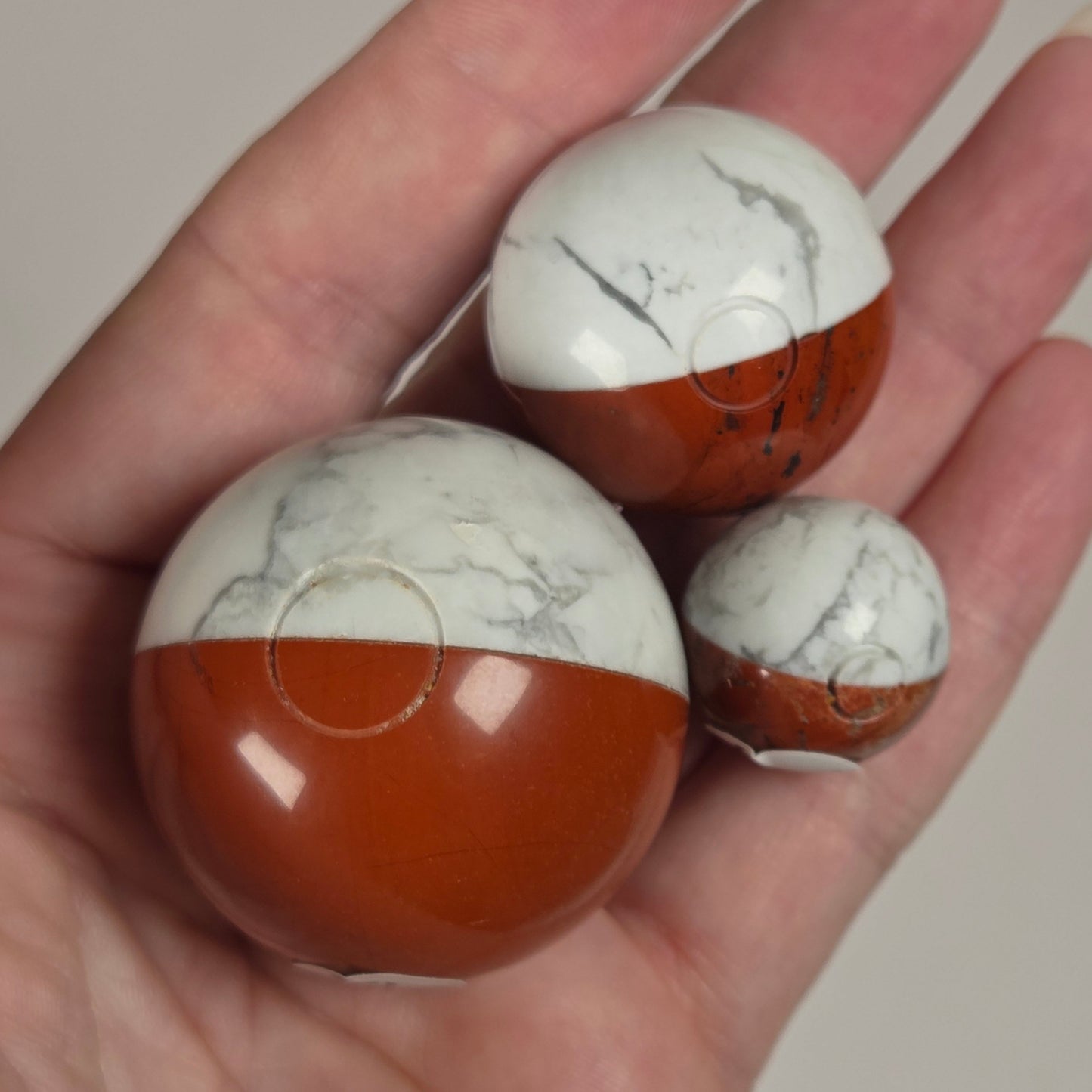 Pokeball carvings made of Howelite and Red Jasper in various sizes being held in a hand against a white background