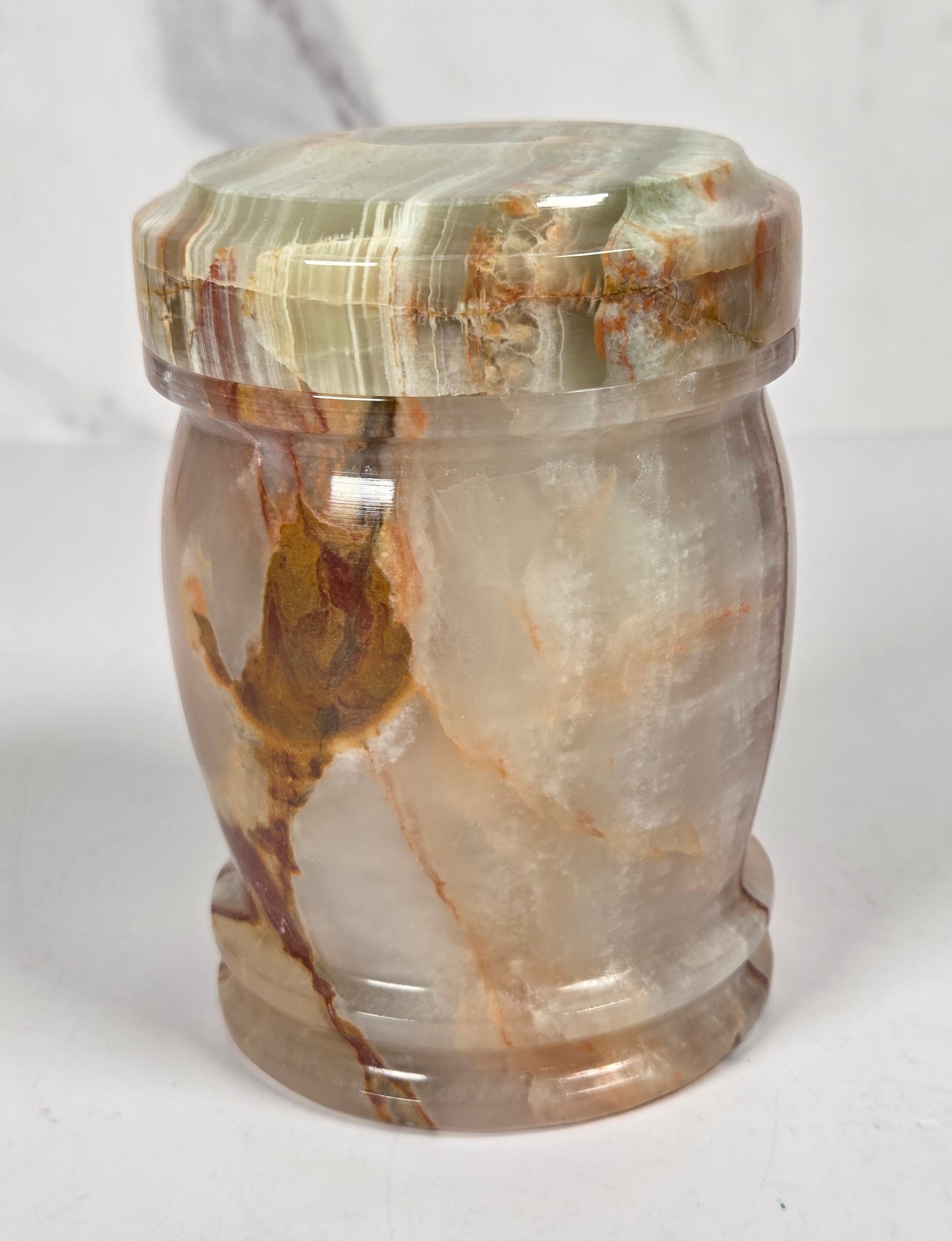 small decorative lidded jar made from polished agate stone, featuring swirling patterns of green, brown, beige, and reddish tones, on a white surface with a white and gray marble background.