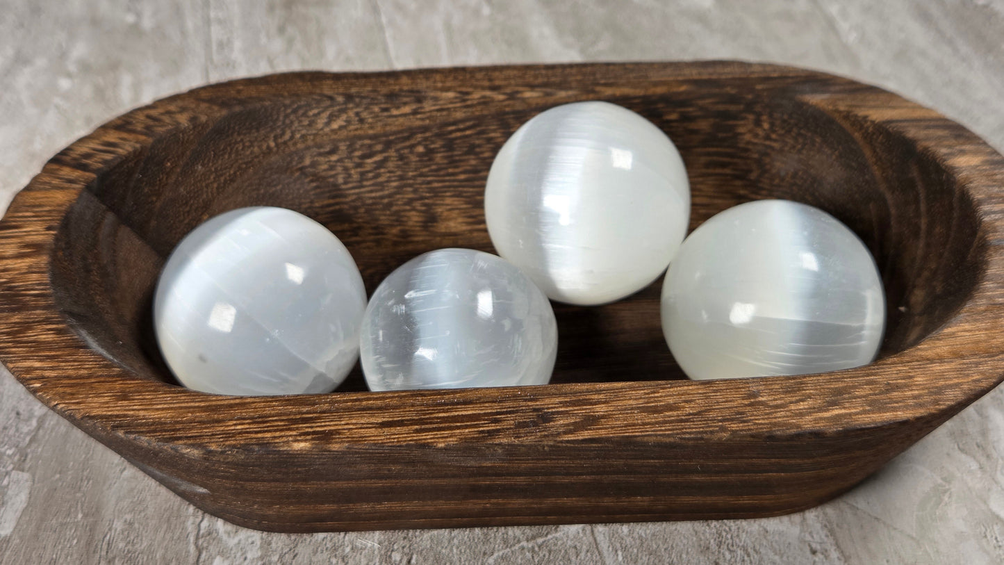 Four white selenite gemstone spheres resting in a dark brown wooden bowl on a textured gray stone surface.
