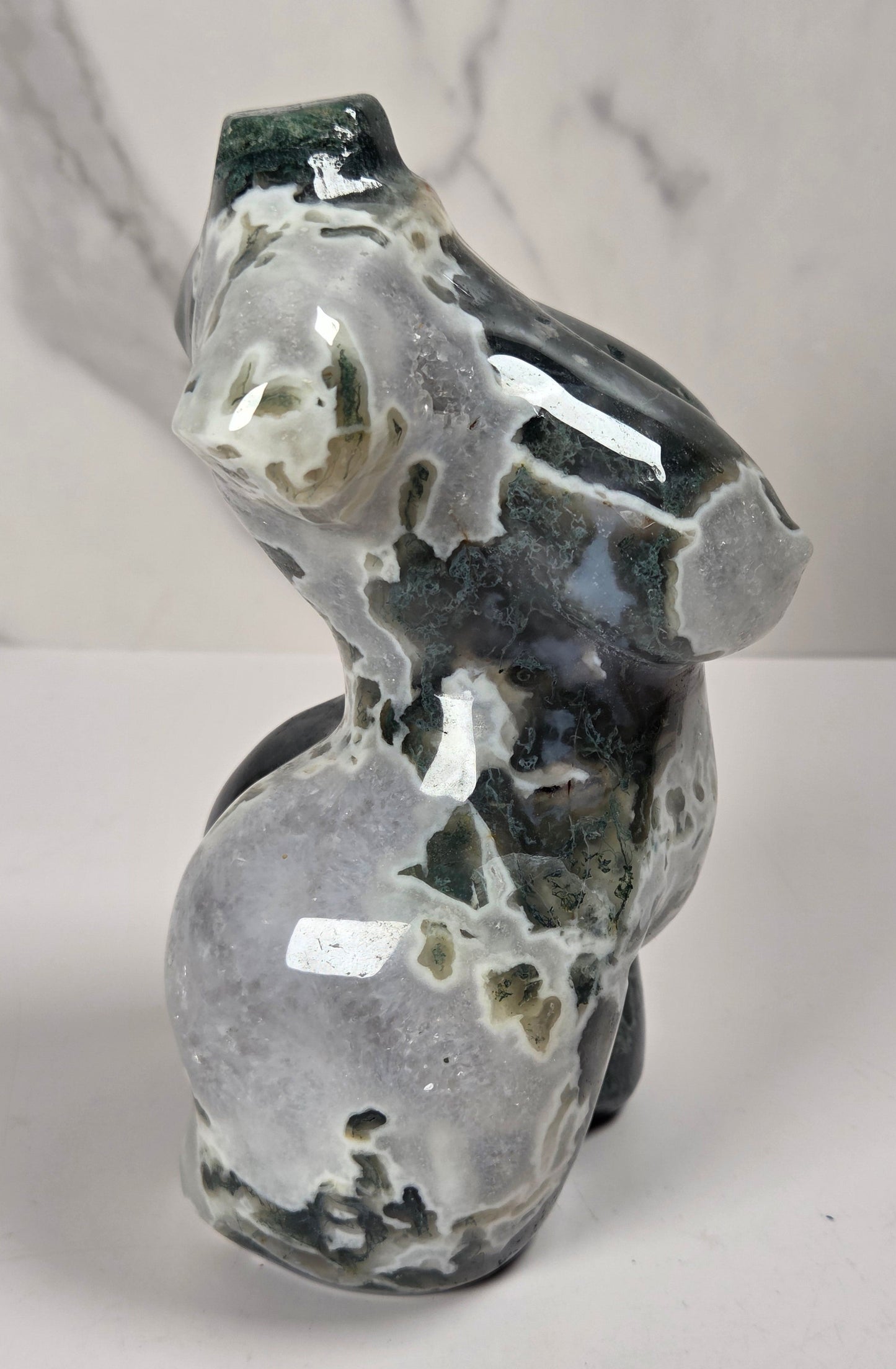 A polished natural stone sculpture shaped like a stylized female torso, shown against a white marble-patterned background.