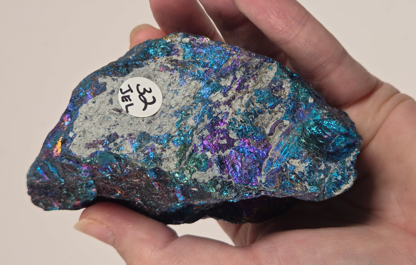 A hand holding an iridescent mineral specimen with shimmering blue, purple, and green tones on a plain beige background. A small white label with handwritten markings is attached to the specimen.