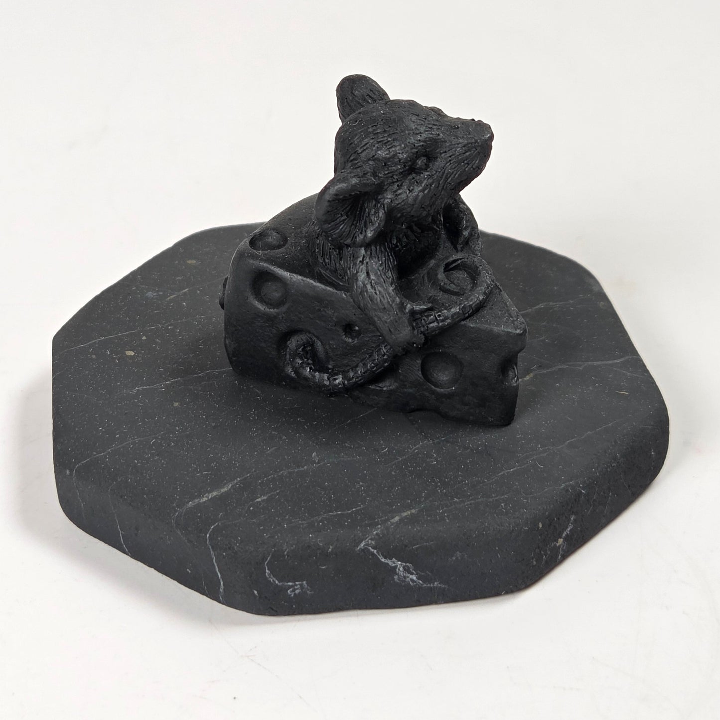 A small black figurine of a mouse sitting on a block of cheese, mounted on a dark gray octagonal base, photographed on a plain white background.