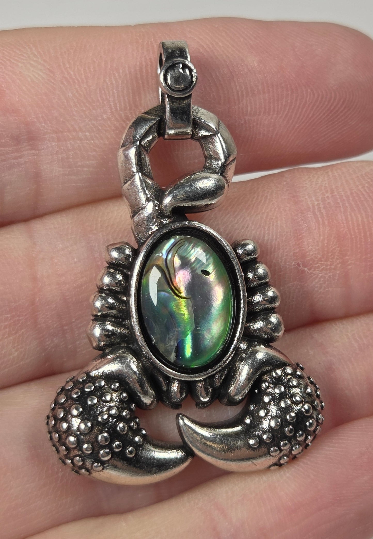 A silver lobster claw pendant with an iridescent abalone shell center, held on a person's fingers against a light gray background.