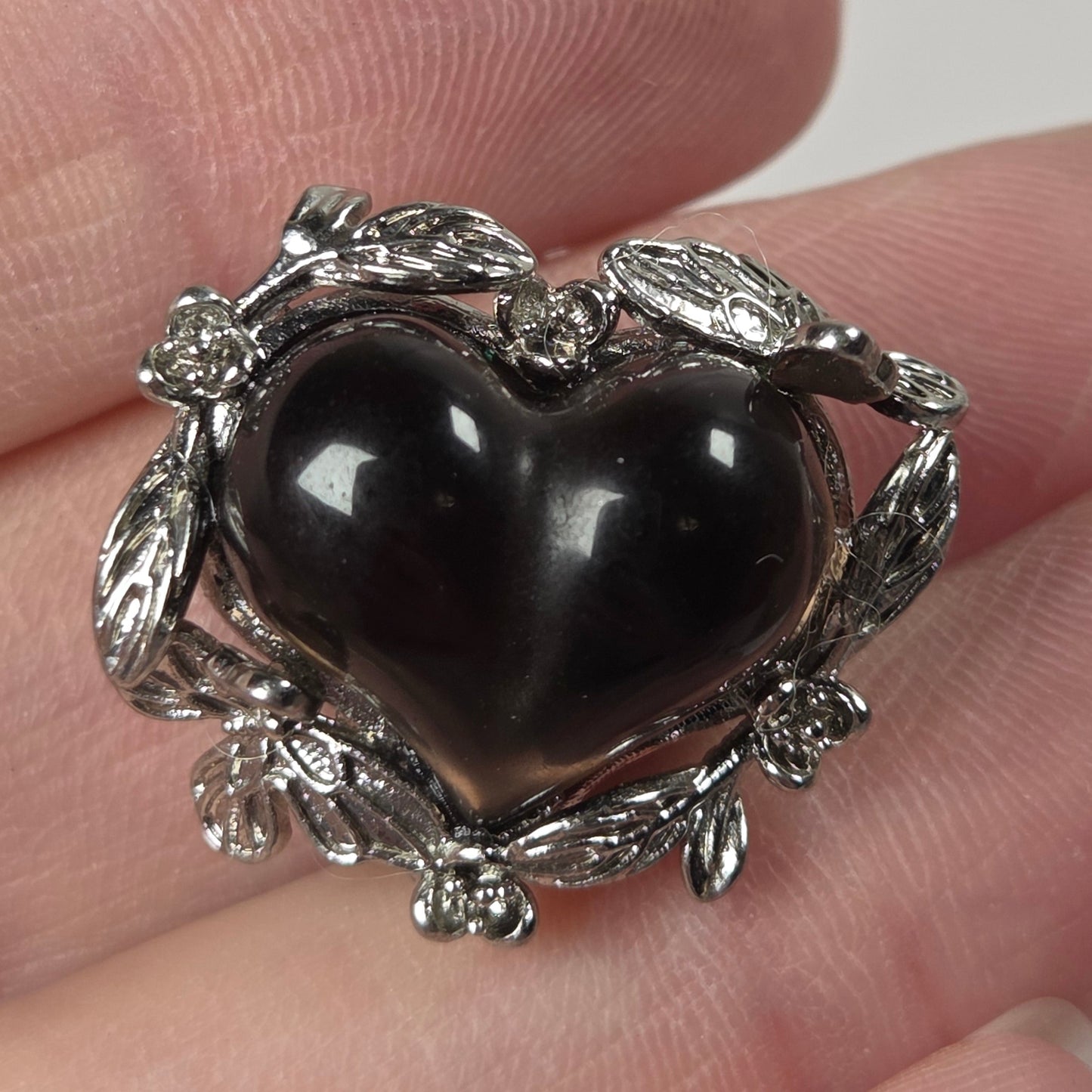 A heart-shaped black stone brooch with a silver leafy border, shown resting on a person's fingers against a neutral light gray background.