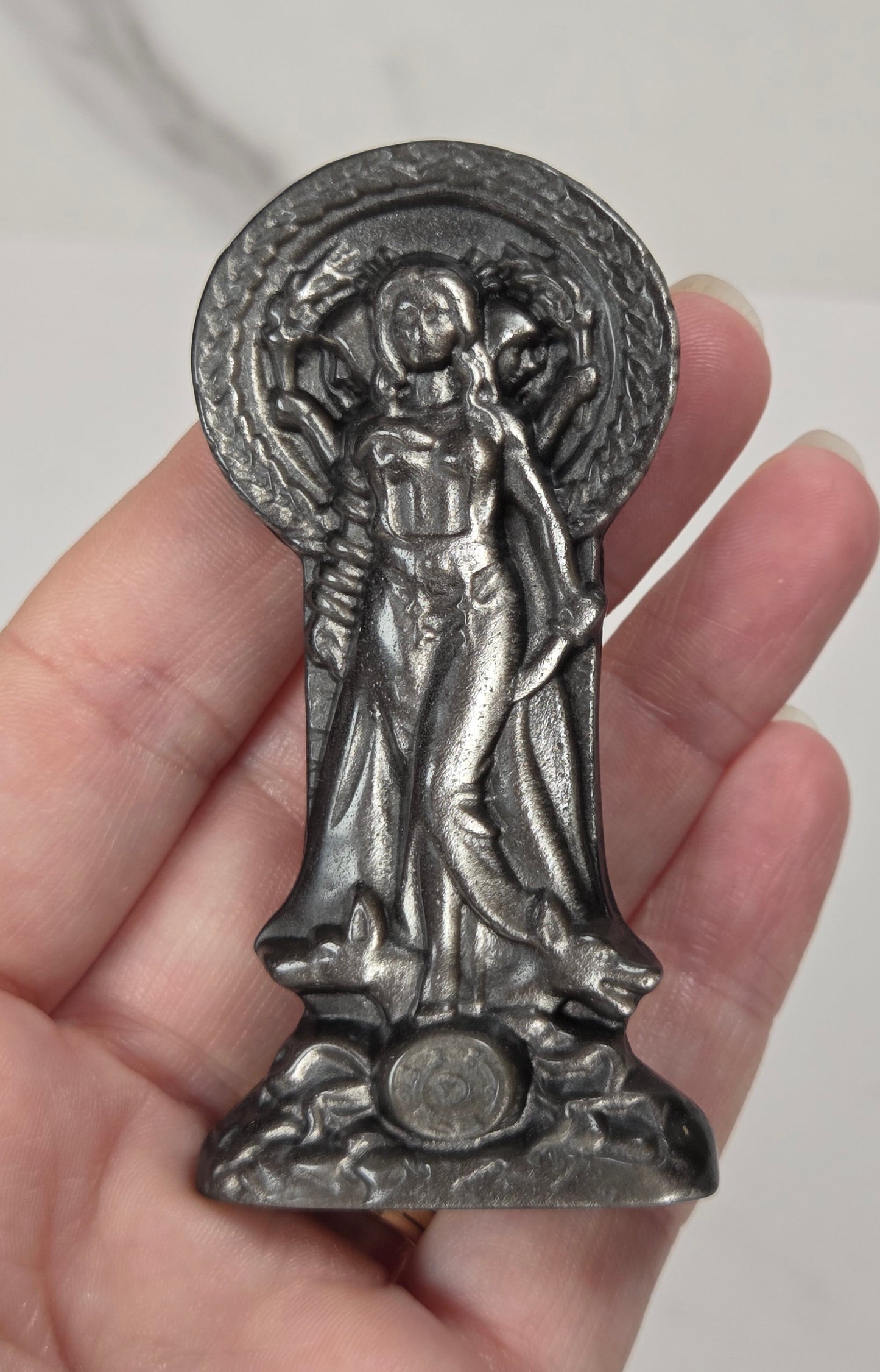 A hand holding a small silver-toned religious figurine of Hecate, shown against a light marble-patterned background.