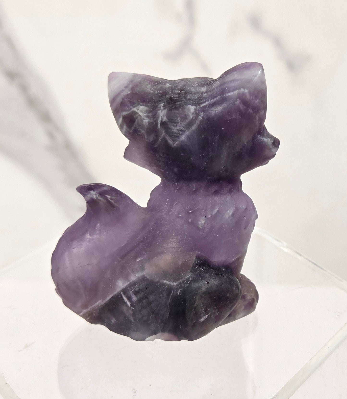 A small carved purple stone figurine shaped like a fox, displayed on a clear acrylic stand against a light marble-patterned background.