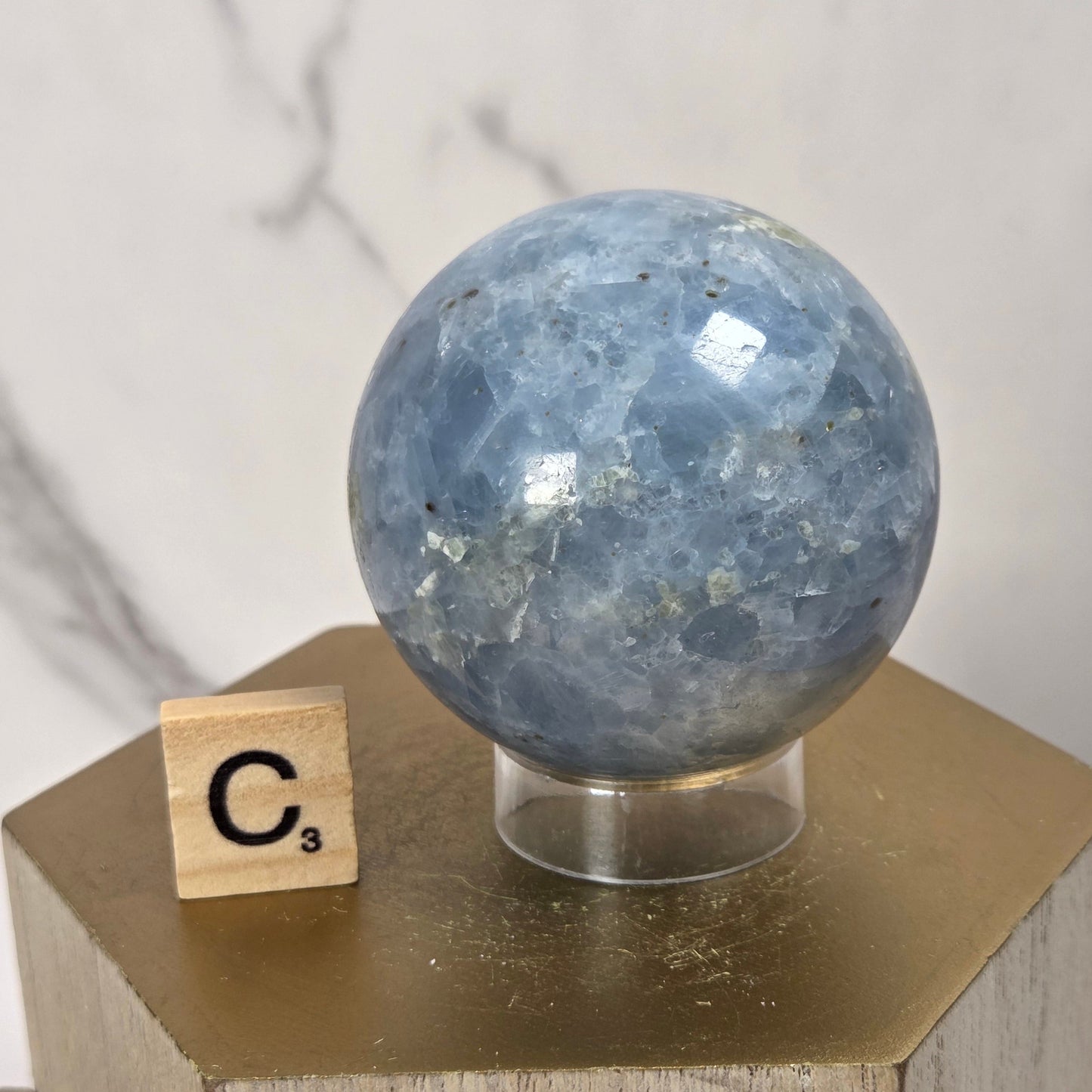 A polished blue stone sphere displayed on a clear stand atop a gold hexagonal pedestal, with a small wooden Scrabble tile showing 'C3' beside it against a light marble-patterned background.