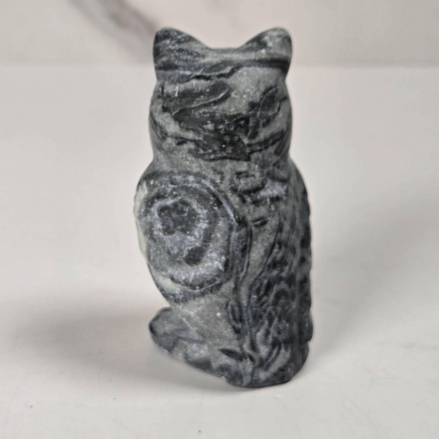 A small carved stone owl figurine with a mottled dark gray and white pattern, standing upright on a plain light surface.