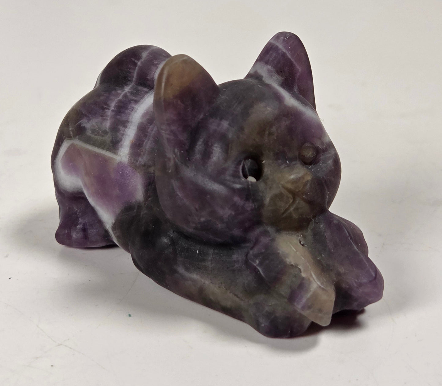A small carved figurine of a cat lying down, made from purple and white marbled stone, set against a plain off-white background.