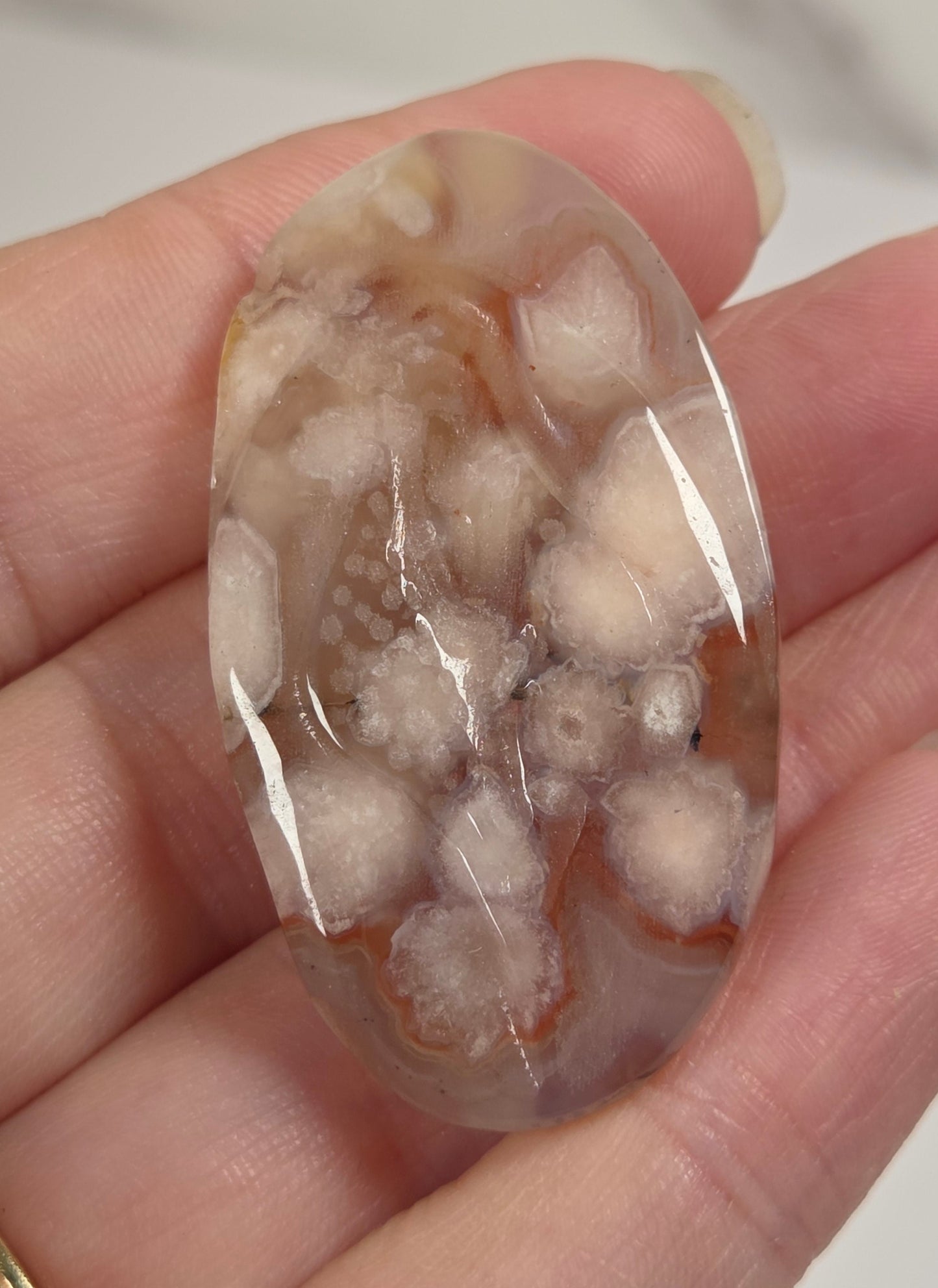 A hand holding a polished oval cabochon gemstone with a marbled pattern of white, beige, and light brown, with a gold band ring visible on the finger.