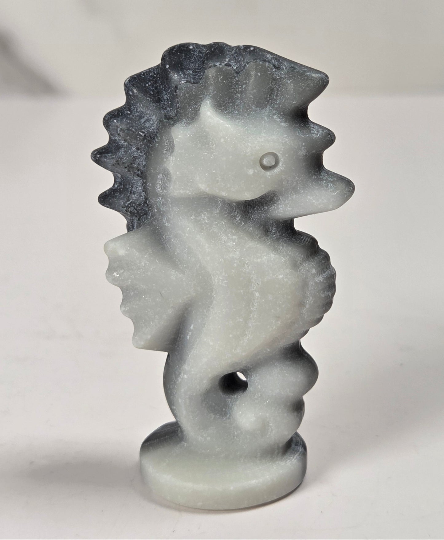 A small carved seahorse figurine in a light gray stone-like material with darker gray shading, standing on a round base against a plain light background.