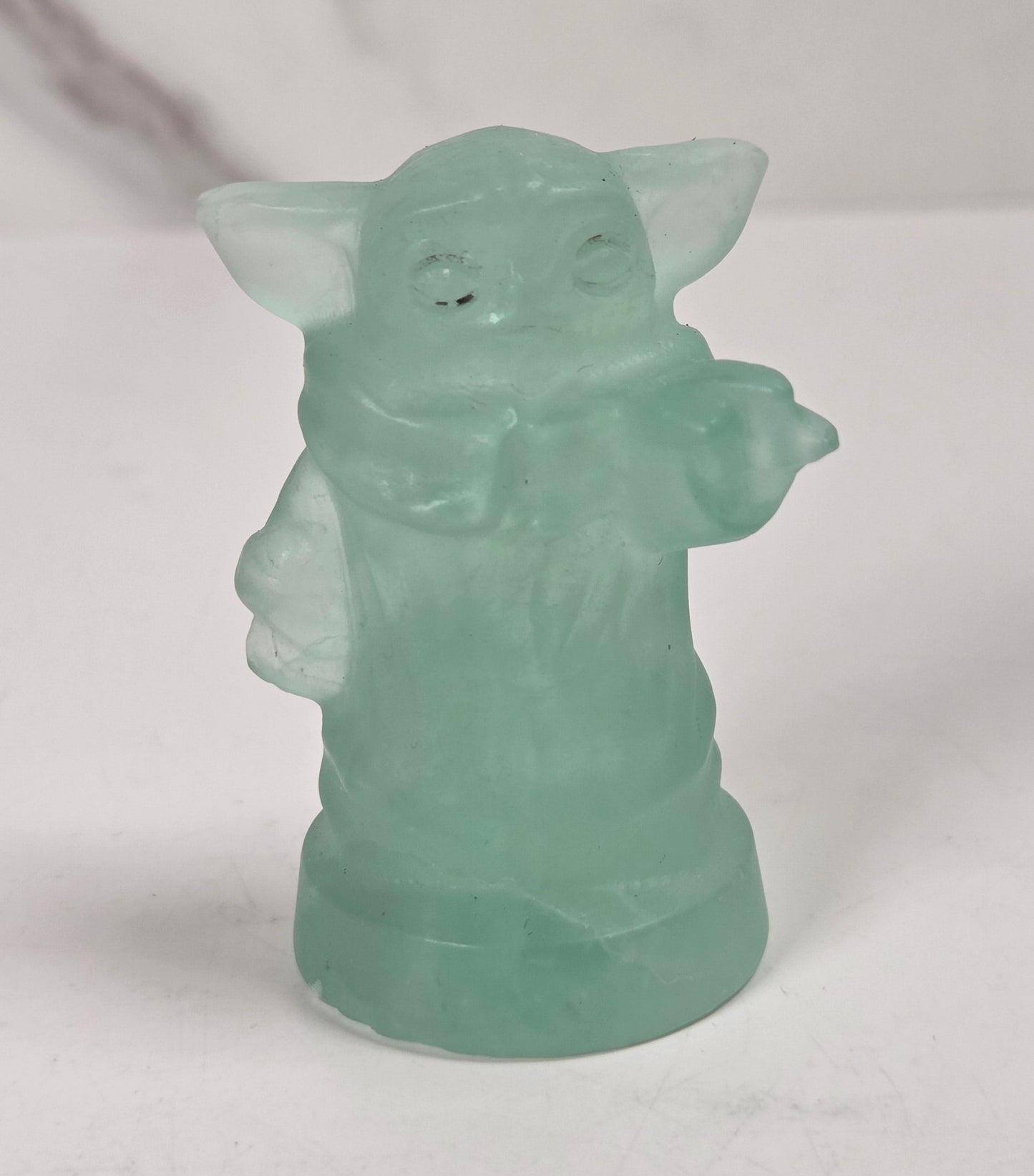 A small translucent mint green figurine of Grogu, standing on a white surface against a light gray and white marble-patterned background.