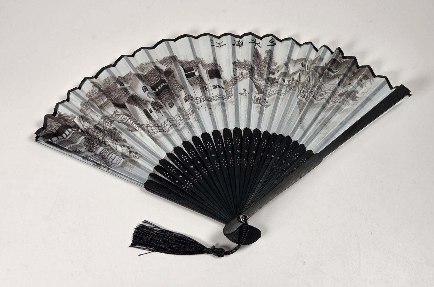 A traditional folding hand fan with black bamboo ribs, a black tassel, and a printed black and white scenic landscape design on the fabric panels, displayed against a plain off-white background.