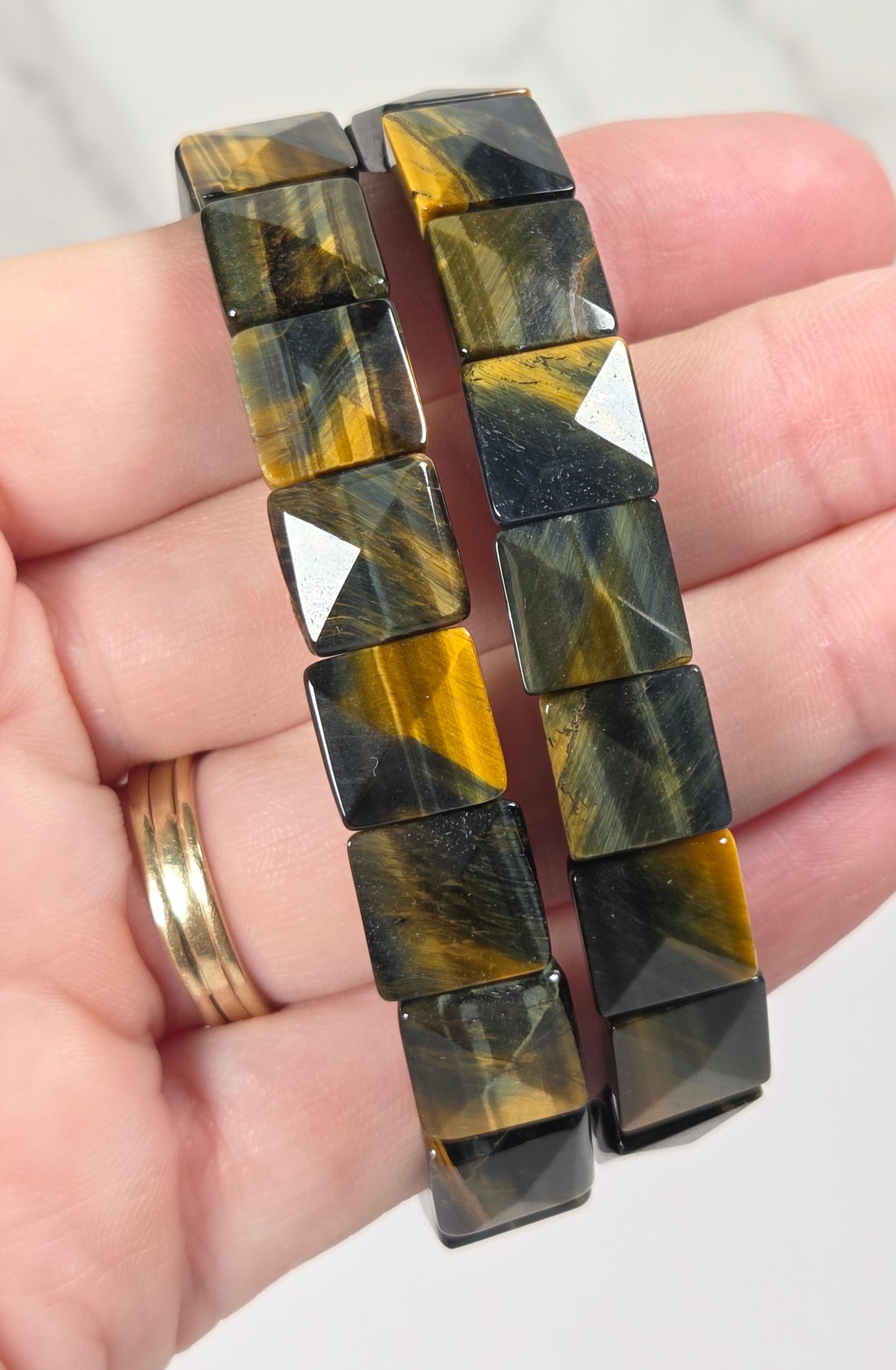A pair of chunky cuff bracelets made of faceted square tiger's eye beads, held in a person's hand. The bracelets feature a dark brown and black base with vibrant golden-yellow striping.