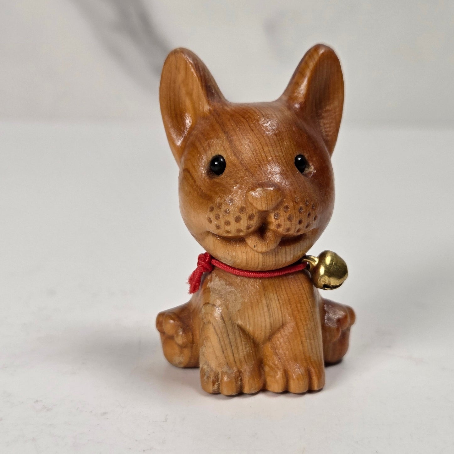 A small carved wooden dog figurine with a red collar and a small gold bell, photographed on a light neutral surface.
