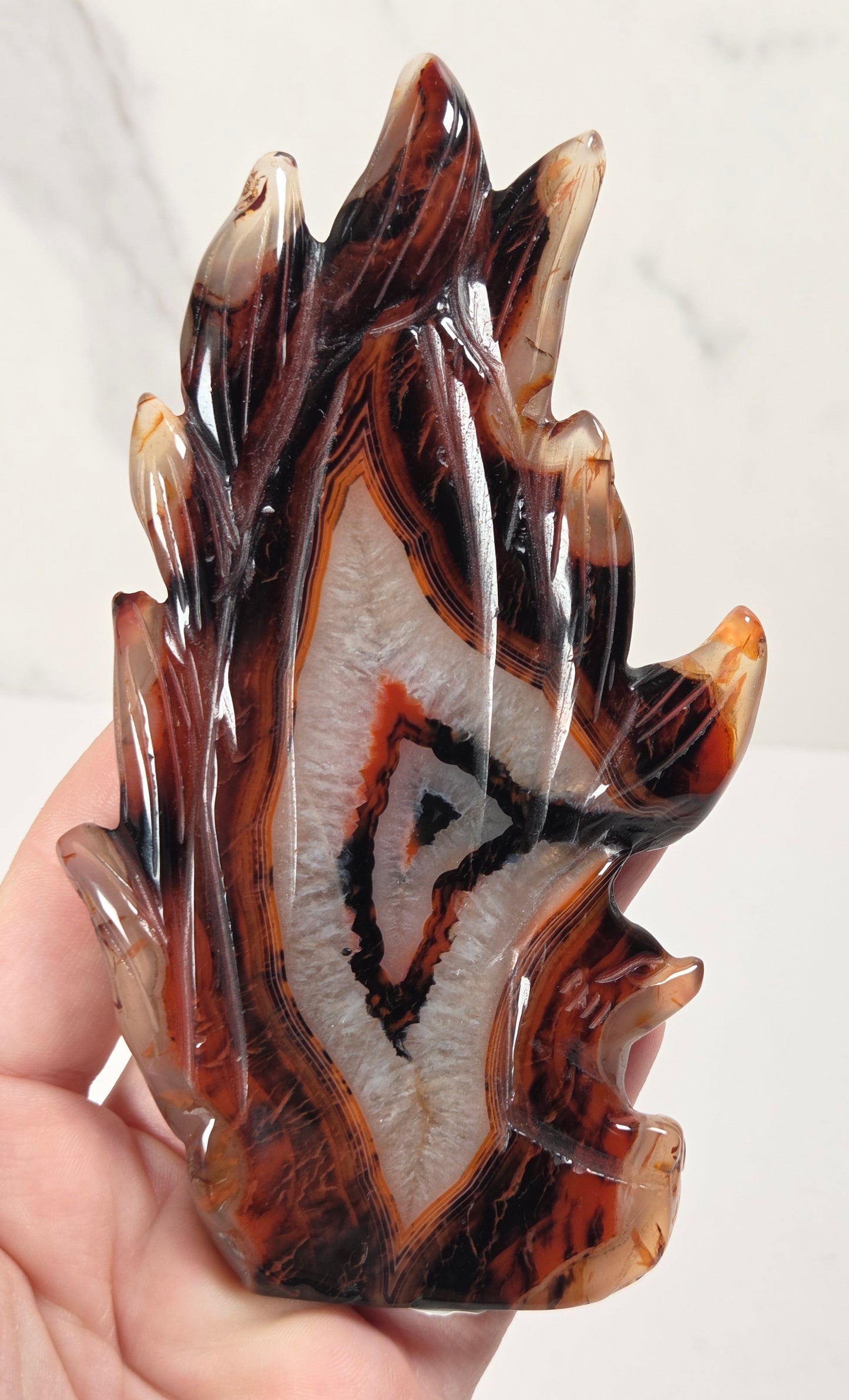 A hand holding a polished agate crystal carving shaped like a stylized flame, showcasing natural banding in amber-brown, black, and creamy white.