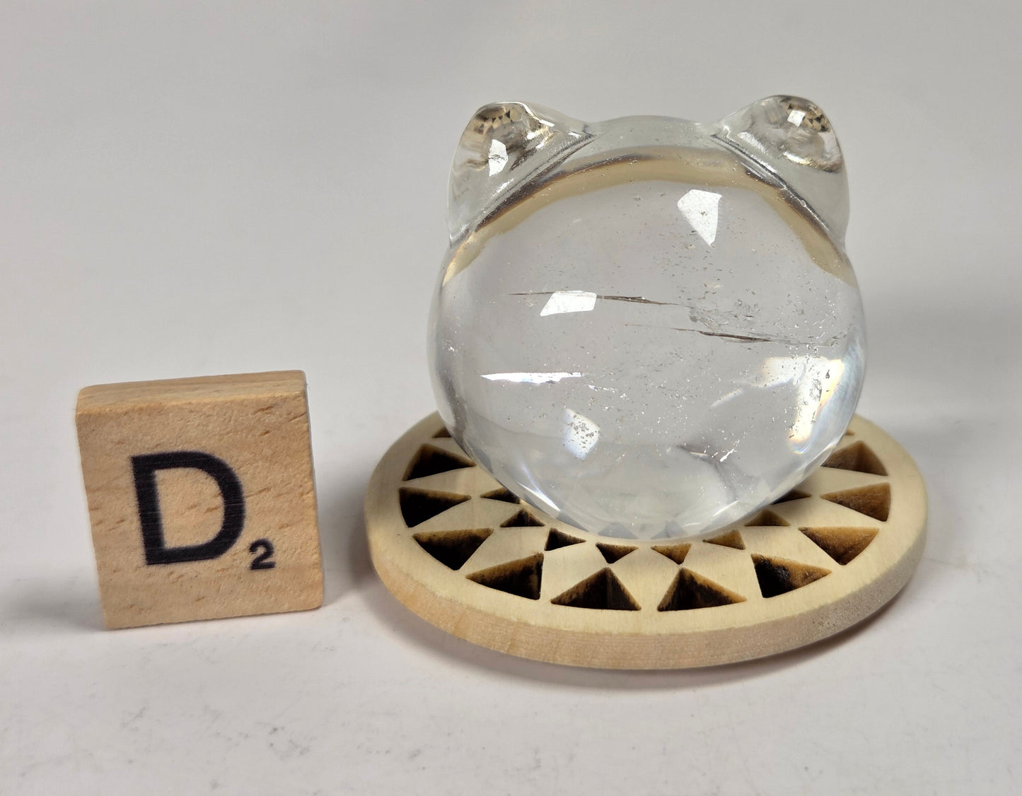 A clear crystal ball with a small frog head cutout at the top, displayed on a round wooden stand next to a wooden Scrabble tile marked D2, on a light gray surface.