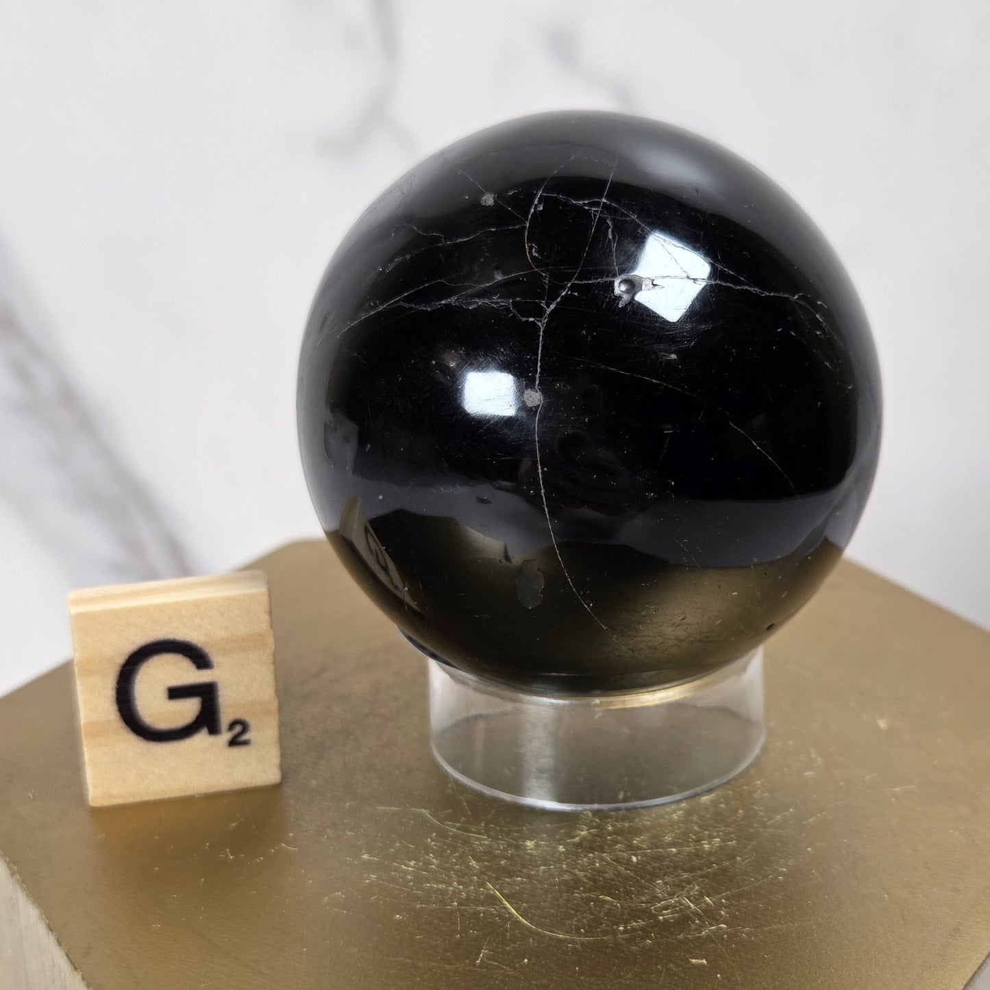 A polished black stone sphere with natural white veining, displayed on a clear acrylic stand next to a wooden Scrabble tile on a gold hexagonal pedestal.
