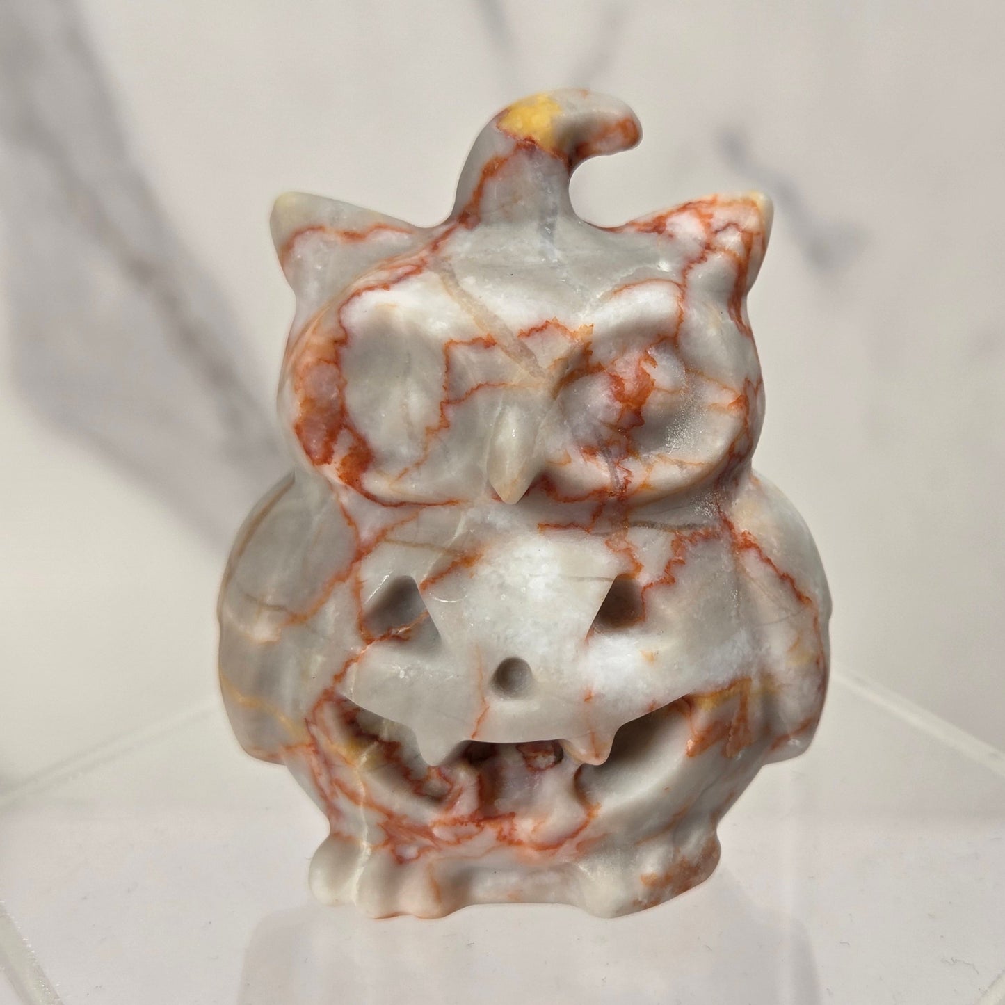 A carved stone owl figurine with orange and brown marbling on a white base, displayed on a clear acrylic stand against a light marble-patterned background.