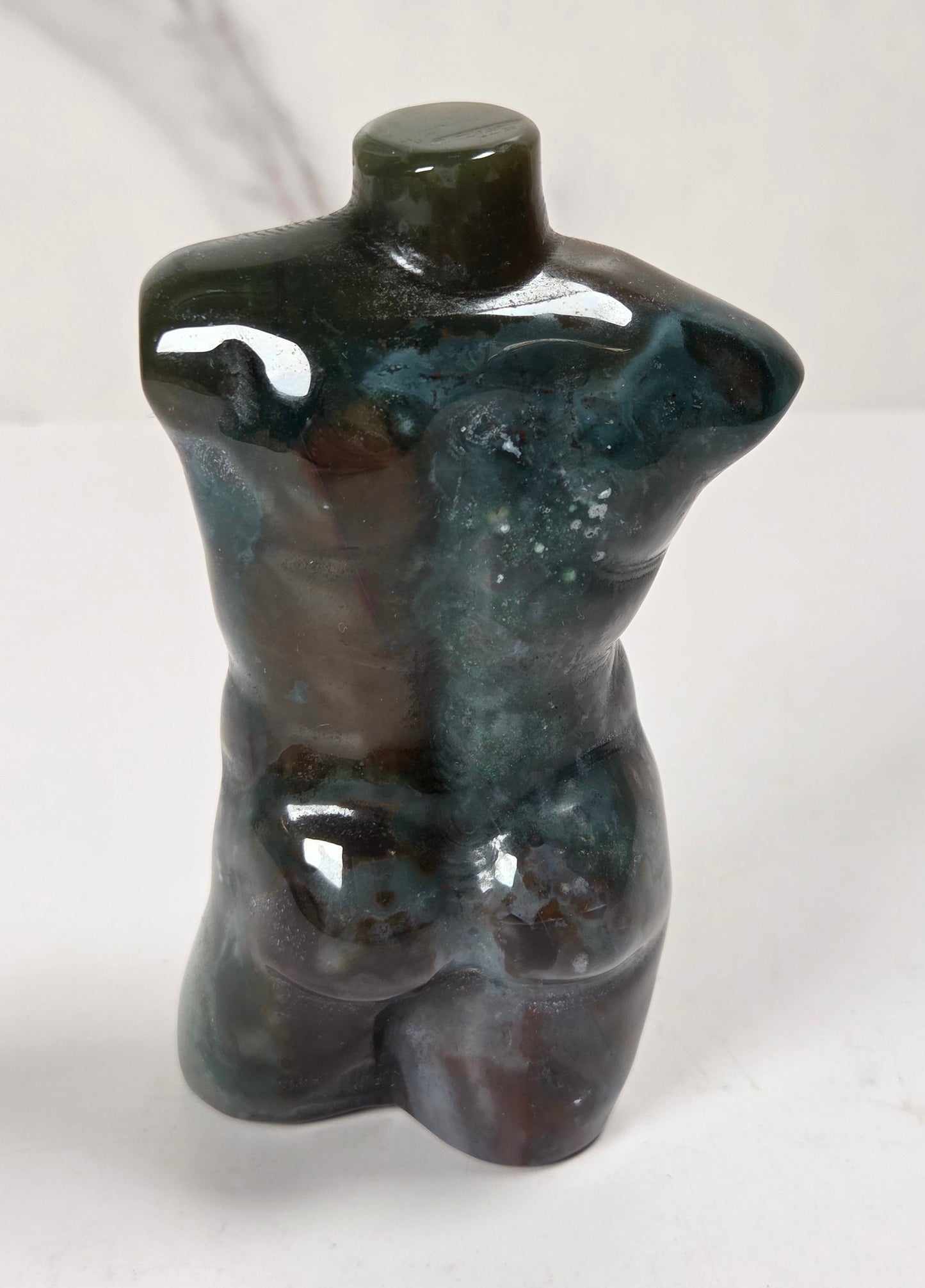 A small glossy torso-shaped decorative sculpture or figurine in dark blue-green and brown marbled tones, photographed against a light marble-patterned background.