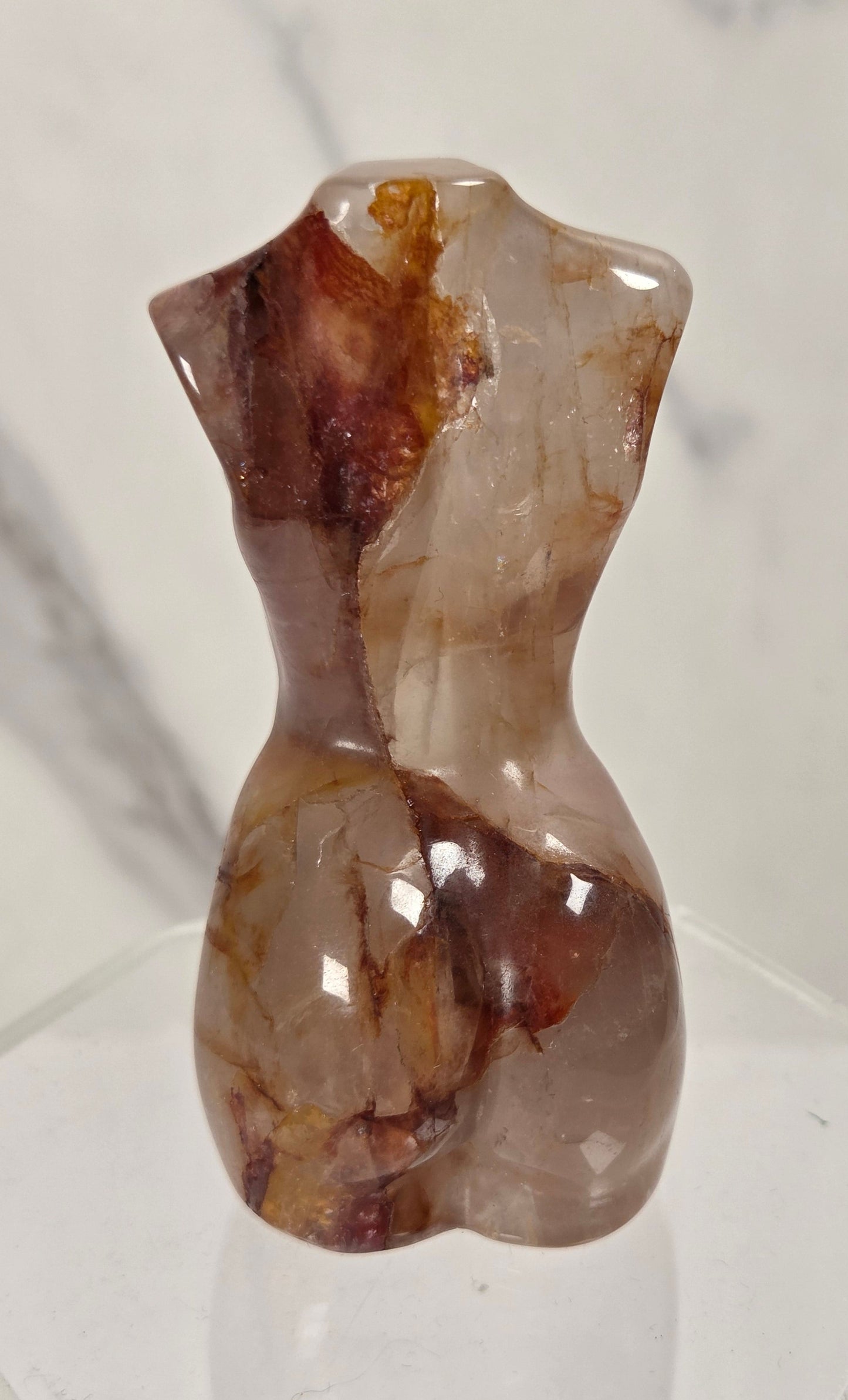 A small polished stone torso sculpture with a glossy finish, featuring natural brown and beige marbling, displayed on a clear acrylic hexagonal base.