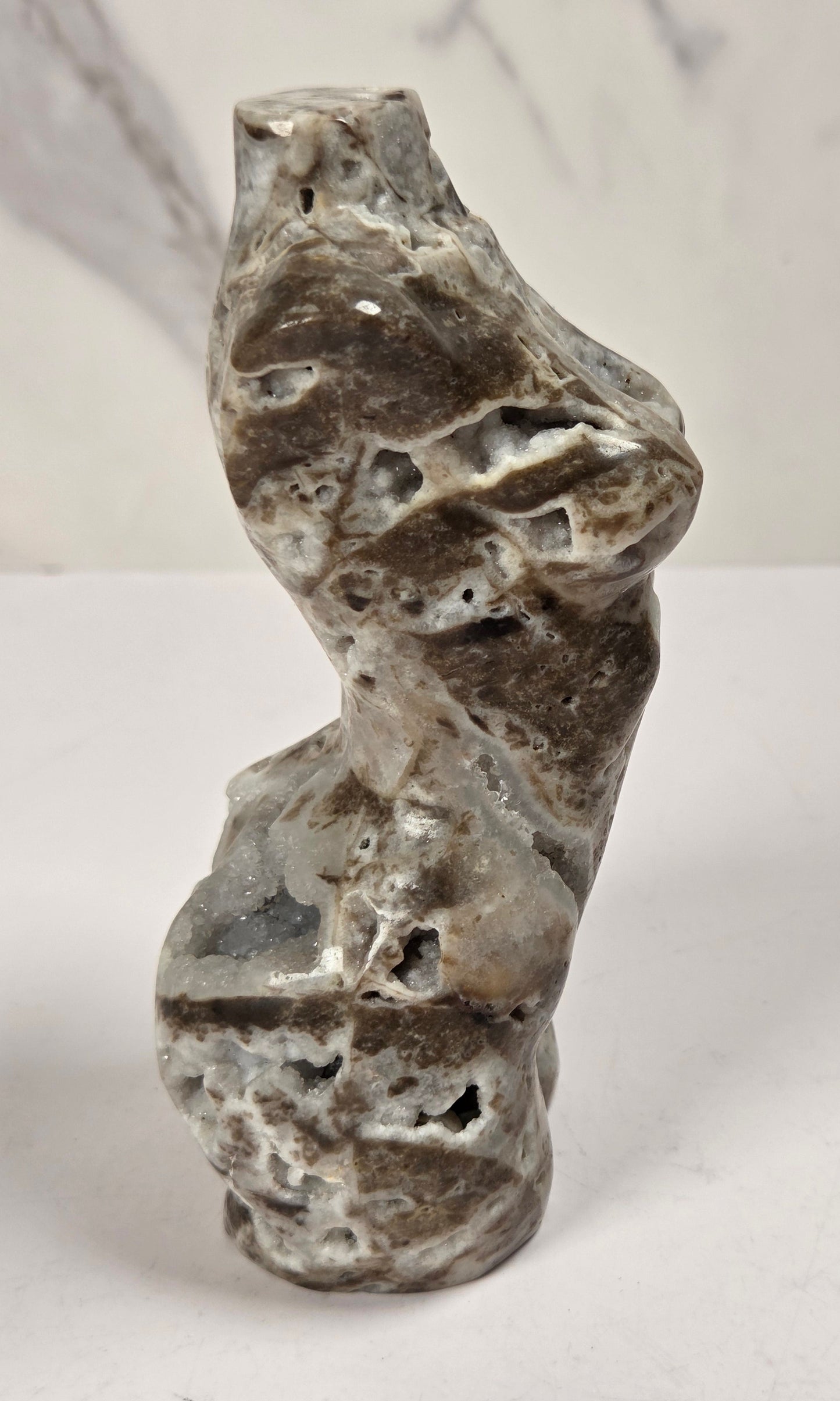 A small sculptural stone figurine with a polished, organic shape, standing upright on a white surface against a light gray marble-patterned background.