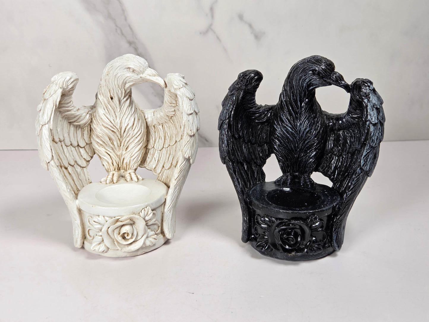Two decorative eagle figurines with spread wings, each mounted on a circular base with floral detailing. One is off-white/ivory and the other is glossy black, displayed side by side on a light surface against a pale marble-pattern backdrop.
