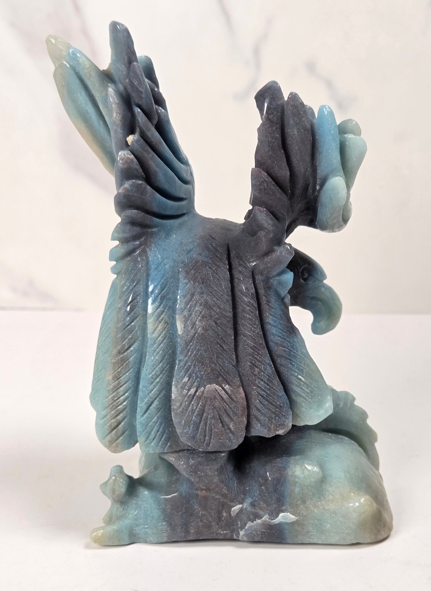 A carved stone sculpture of a bird with wings spread, featuring shades of blue, gray, and teal, set against a light marble-patterned background.